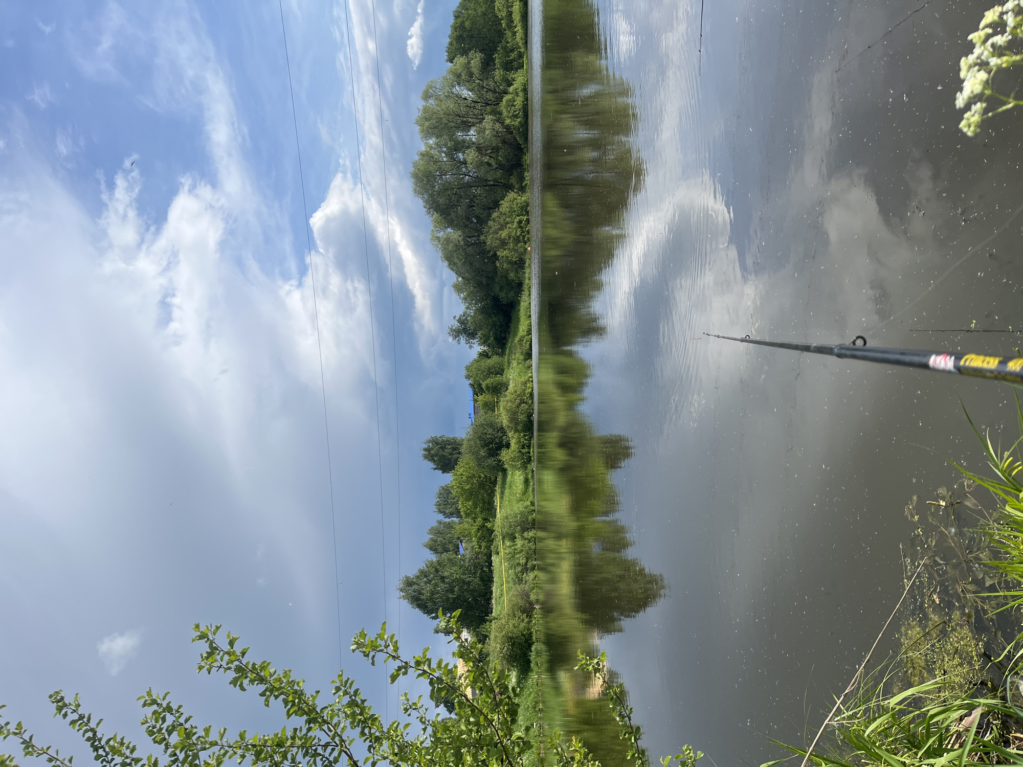 The image presents a riverside scene in Фоки, Russia. A body of water, presumably a river or lake, reflects the sky and surrounding trees, creating a mirrored effect. Vegetation lines the banks, with trees dominating the landscape. A fishing rod is visible, extending into the water from the right side of the frame. Power lines stretch across the sky. The sky shows a mix of clear blue and cloudy areas.