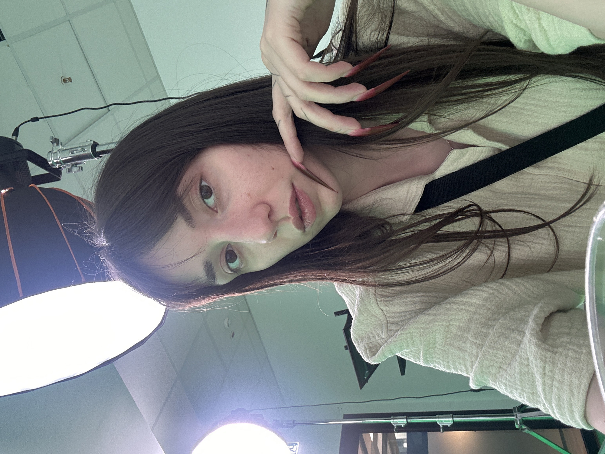 A woman with long, dark hair is in a room with studio lighting equipment. She is wearing a light-colored shirt with a black strap across her torso. Her nails are long and painted red. The background includes a ceiling with fixtures and equipment. The location is likely a photography or video studio, and there is no indication of being in Belgrade, Serbia.
