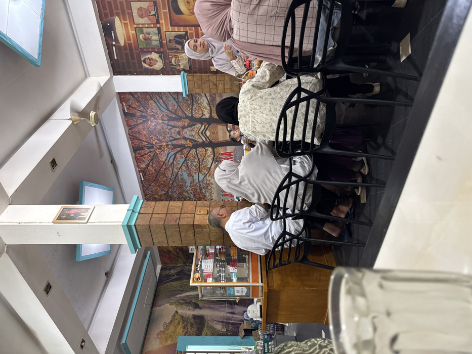 An indoor photograph depicts a restaurant or eatery located in Payameuneng, Indonesia, viewed from a foreground table. In the mid-ground, approximately seven individuals are gathered around a rectangular table. Five adults are seated on black plastic chairs with open backrests. From left to right, a male in a white collared shirt with glasses faces right, his right hand near his face; a female in a light grey hijab faces right; a female in a black hijab faces right; a female in a light patterned long-sleeved shirt has her back to the viewer; and a female in a pinkish-brown checkered top has her back to the viewer. A small child is partially visible between two seated adults. A female in a light grey hijab and light-colored top stands to the right of the table, facing and interacting with the seated group. The table holds several glasses and bottles.
The ceiling is white with recessed lighting and a cream-colored ceiling fan is visible in the upper right. Two large support pillars are present: a central pillar is covered in brown brick-like tiles with light blue decorative accents at the top and bottom, and a framed picture hangs on its side; another white pillar with light blue accents is visible on the far right. The walls feature diverse murals: the left wall displays a waterfall scene with green foliage, while the back wall has a symmetrical autumnal tree scene with red-orange leaves. The right wall and parts of the back wall above the seated group are covered in a brick-patterned wallpaper adorned with multiple framed portraits of individuals. To the far left, a light-colored wooden counter supports a glass display cabinet containing packaged goods, including cigarettes and snacks, with shelves below holding bottled beverages. The floor is dark-colored. A partially visible clear glass sits on the foreground table.