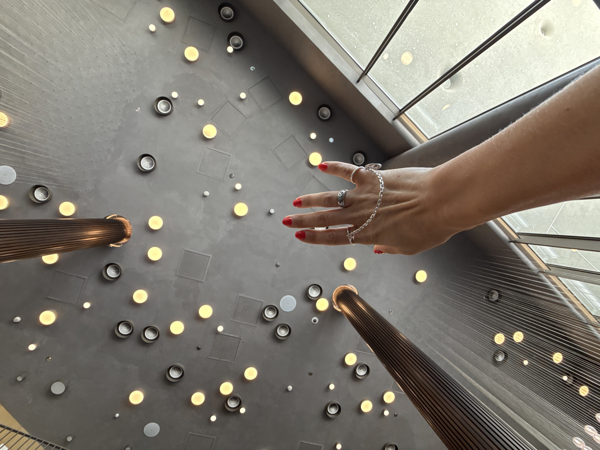 The image shows a hand with bright red nail polish extended upwards towards a modern ceiling. The ceiling is dark gray and features a grid of recessed circular lights, some of which are illuminated with a warm yellow glow. There are also several square ceiling panels visible. A large, ribbed, wooden structure extends vertically from the lower left of the frame, its texture contrasting with the smooth ceiling. A delicate silver chain bracelet with charms is worn on the wrist and extends across the fingers, with rings on two fingers. In the upper right and far right of the image, a large window with metal mullions is visible, with a blurred, reflective outdoor scene beyond. The lighting suggests an indoor setting, possibly a contemporary public space like a shopping mall or an office building. The city of Голубинское сельское поселение, Ukraine, is not depicted or suggested in this image.