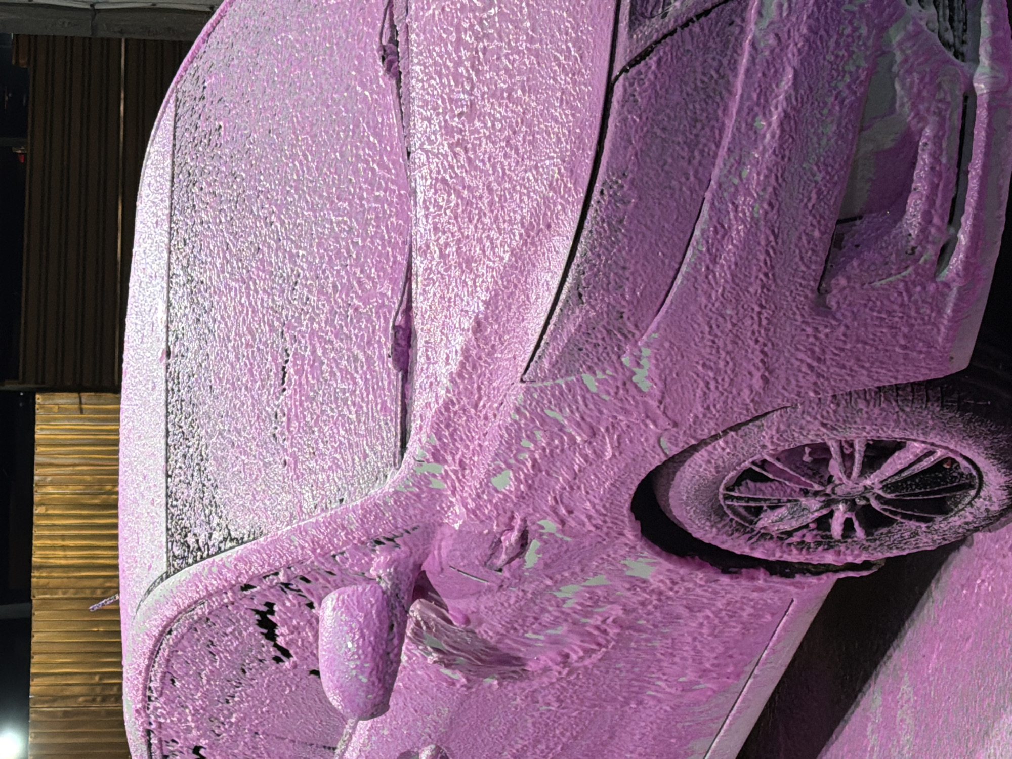 The image shows a pink-foamed car parked on a street, likely at a car wash. The car is a modern sedan, with its windows, hood, and sides completely covered in pink foam. The tires and wheel rims are also partially covered. In the background, a wooden fence is visible, with some structures behind it. The lighting suggests it is either dusk or an indoor setting with artificial lights. The scene appears to be in an urban or suburban area. The location is identified as Korsuntsi, Ukraine. There are no people visible in the image.