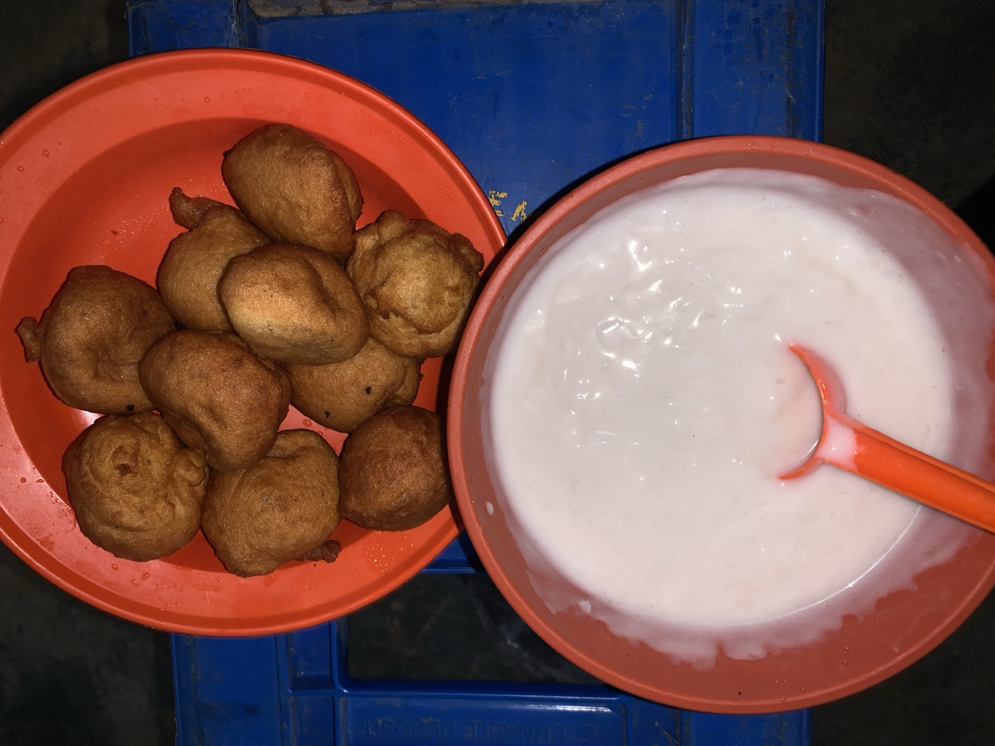 The image displays a top-down view of two coral-red plastic bowls, each containing food, resting on a blue plastic crate.
The bowl on the left is filled with approximately a dozen golden-brown, irregularly shaped fried dough balls, which resemble Nigerian puff-puff. These dough balls vary slightly in size and shape, with a crispy-looking exterior. Some small dark specks are visible on a few of the fried items.
The bowl on the right contains a thick, white, semi-liquid substance, consistent with a creamy porridge, gruel, or a sweet sauce. An orange plastic spoon is partially submerged in this white substance, resting against the rim of the bowl.
The bowls are placed on a blue plastic crate or stool, which appears worn with visible scratches and some dirt. Faint, inverted white text is present on the side of the blue crate. Though largely indistinct, fragments like "BETULEN" and "DEPOST" or "DEPOSE" are partially visible, suggesting a label or brand name, possibly in French or another language.
The lighting in the image is dim, indicating an indoor setting or perhaps nighttime. There are no people visible, and the scene depicts a simple arrangement of food ready for consumption. The image cues suggest a casual eating environment, possibly at a market stall, street vendor, or a home in New Stadium, Nigeria.