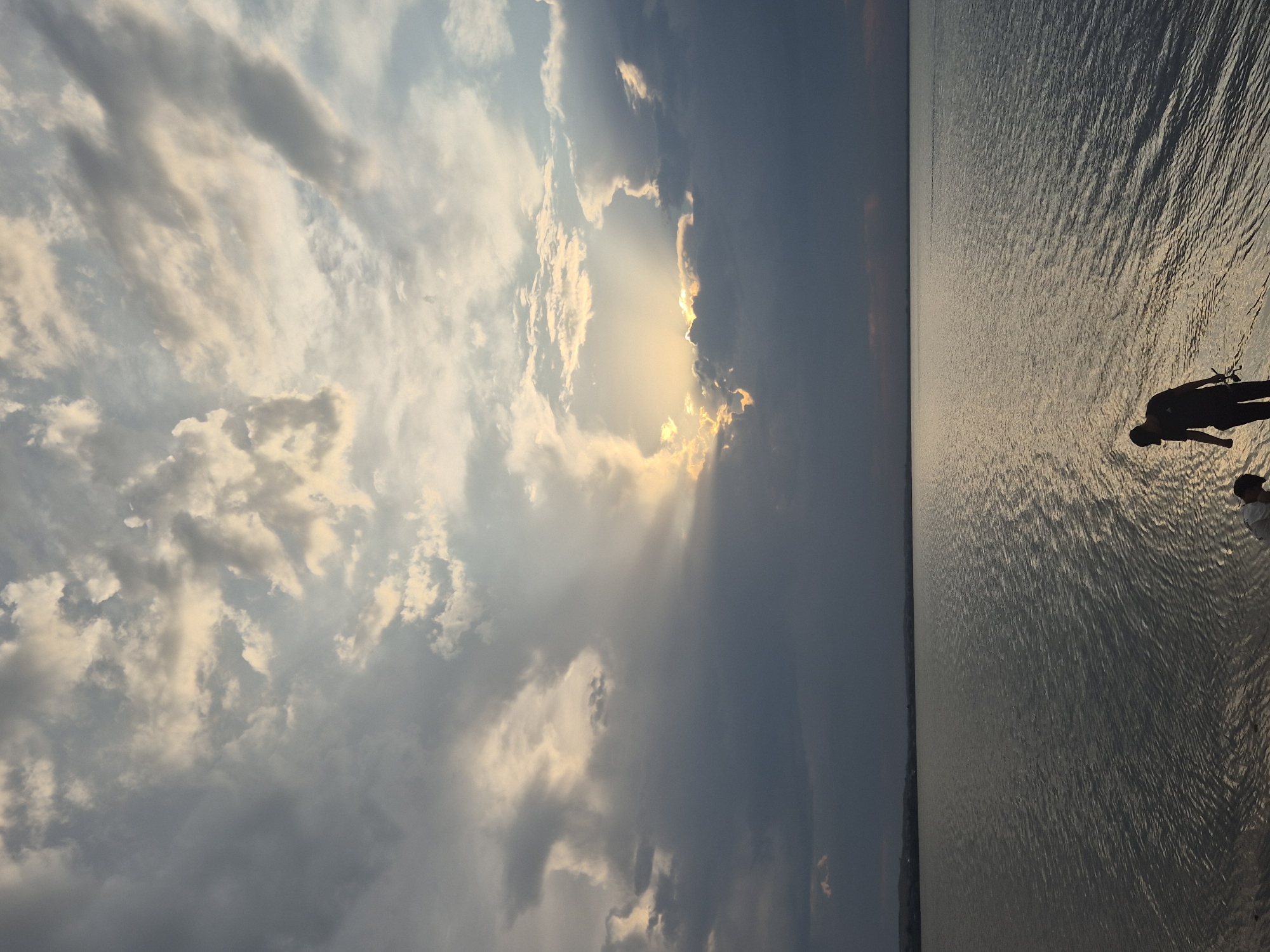 An outdoor, eye-level shot captures a serene seascape under a dramatic, cloudy sky. The water, rippling gently, reflects the muted light of the setting sun, which peeks through a break in the clouds, casting a golden glow. On the sandy shore in the foreground, two figures are partially visible. One, wearing a dark t-shirt and cap, stands facing the water, holding what appears to be a fishing rod. Another person, with dark hair and wearing a white shirt, is partially visible to the left of the first figure, crouched down. In the distance, a faint silhouette of a shoreline with hills is visible. The overall scene suggests a peaceful evening by the water, possibly Lake Burabay, a prominent natural landmark near Kokshetau, Kazakhstan.