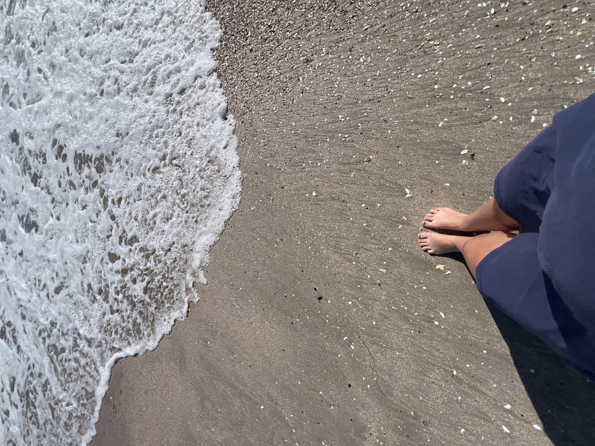 The image captures a low-angle view of a person's feet standing on a sandy beach with small shells. The edge of a foamy wave is encroaching on the sand from the top of the frame. The scene is likely located on the coast of Pomorie, Bulgaria, a city known for its beaches and mineral salt lakes. The sand is damp and appears to be mixed with a significant amount of shell fragments. The person is wearing navy blue shorts or a skirt. Their toenails are painted dark purple. A shadow is cast to the left of the person's legs, indicating a light source, likely the sun, from the right. The overall environment suggests a sunny day at the beach.