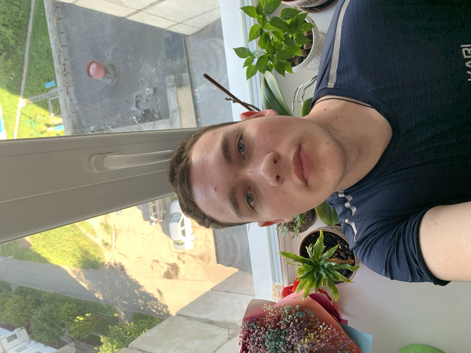 A young man is captured in a selfie, positioned indoors by a window that overlooks an urban or suburban outdoor scene. He has light eyes, short dark hair, and visible stubble, wearing a dark blue ribbed t-shirt with a light grey stripe on the sleeve and partial white text that appears to be "KARI." A thin black string or necklace is visible around his neck.
Through the window, a bright, sunny daytime environment is visible, with strong sunlight creating clear shadows. The view includes concrete paths, green grassy areas, and trees. A road with a white car is visible, and further in the distance, what looks like a children's playground with blue and green equipment can be seen. Below, on a paved surface, a pink or red bucket-like object is situated.
On the windowsill beside the man, several potted plants are arranged, including one with large green leaves and another smaller, spiky plant. An orchid stem is also present. In the foreground, a colorful bouquet of flowers rests on the sill, featuring vibrant, multicolored baby's breath and a few larger blossoms, wrapped in pink and light blue paper. The setting appears to be an apartment in a location such as Severodvinsk, Russia.