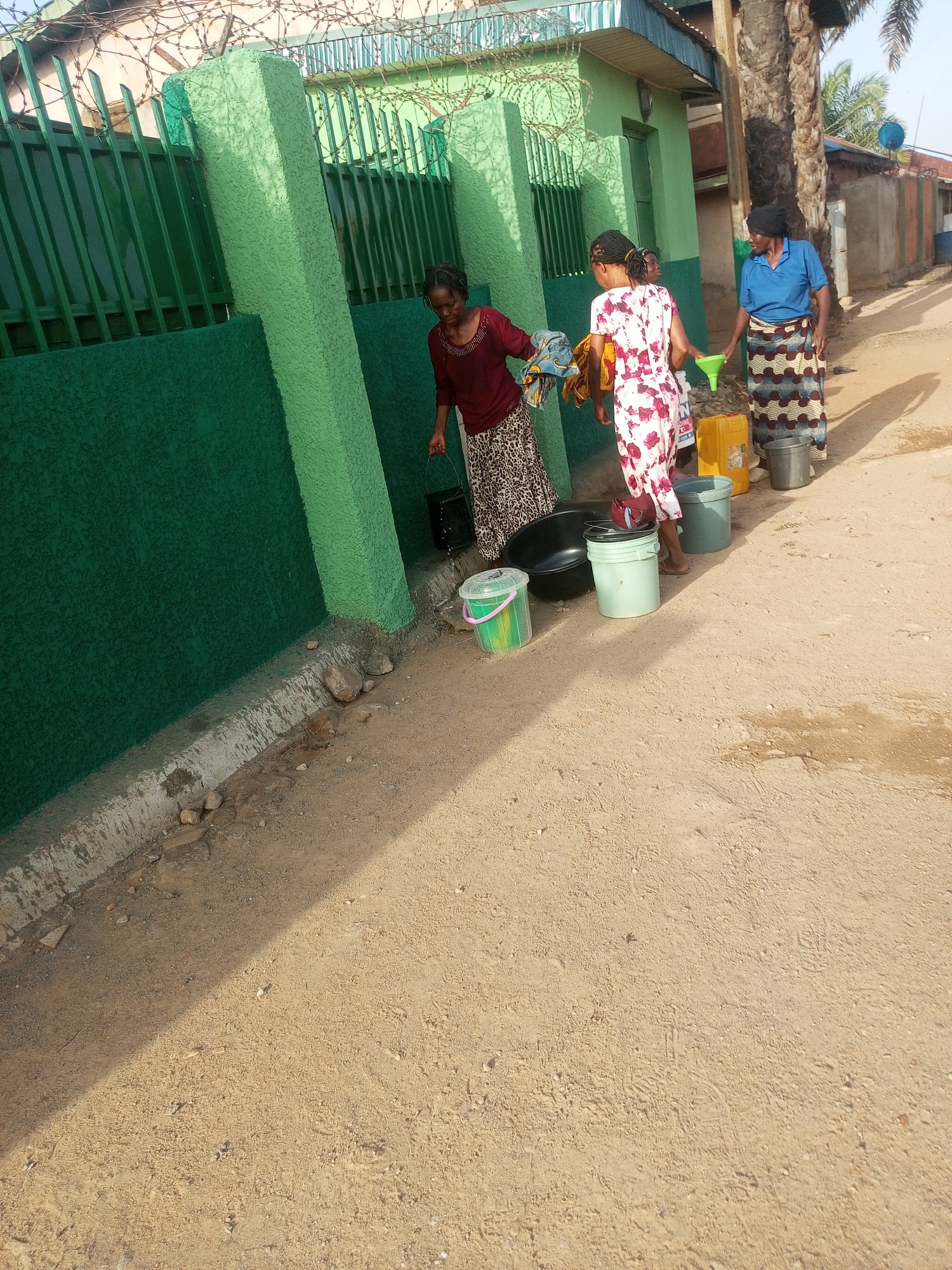 This media file captures an outdoor scene in Sabon Tasha, Nigeria, featuring three women engaged in a water-related activity alongside a green building and fence.
In the foreground, a light brown, unpaved dirt road or pathway dominates the lower half of the image. On the left, a vibrant green wall and fence, topped with barbed wire, run along the side of the road.
Three women are visible. On the left, a woman wearing a dark red top and a patterned skirt is holding a black bucket, seemingly pouring water from it into a lighter green bucket on the ground. She appears focused on her task. In the center, another woman in a white and floral patterned dress is holding folded clothes and is positioned near a large black basin and a light green bucket. Her back is mostly to the viewer. To her right, a third woman, dressed in a blue top and a patterned skirt, holds a green funnel over a yellow jerrycan, indicating she is either filling or emptying the jerrycan. Several other buckets of various colors (grey, clear with pink accents) are scattered around the women on the ground.
The activity suggests the collection, transfer, or use of water, possibly for domestic chores like washing or drinking. The bright lighting indicates it is daytime, likely mid-morning or early afternoon, given the clear shadows cast by the people and objects. Behind the women, part of a building with a light green exterior and a brown roof is visible, along with palm trees and other structures further down the road, consistent with a community setting. The yellow jerrycan has some faint, unreadable text on its side.
