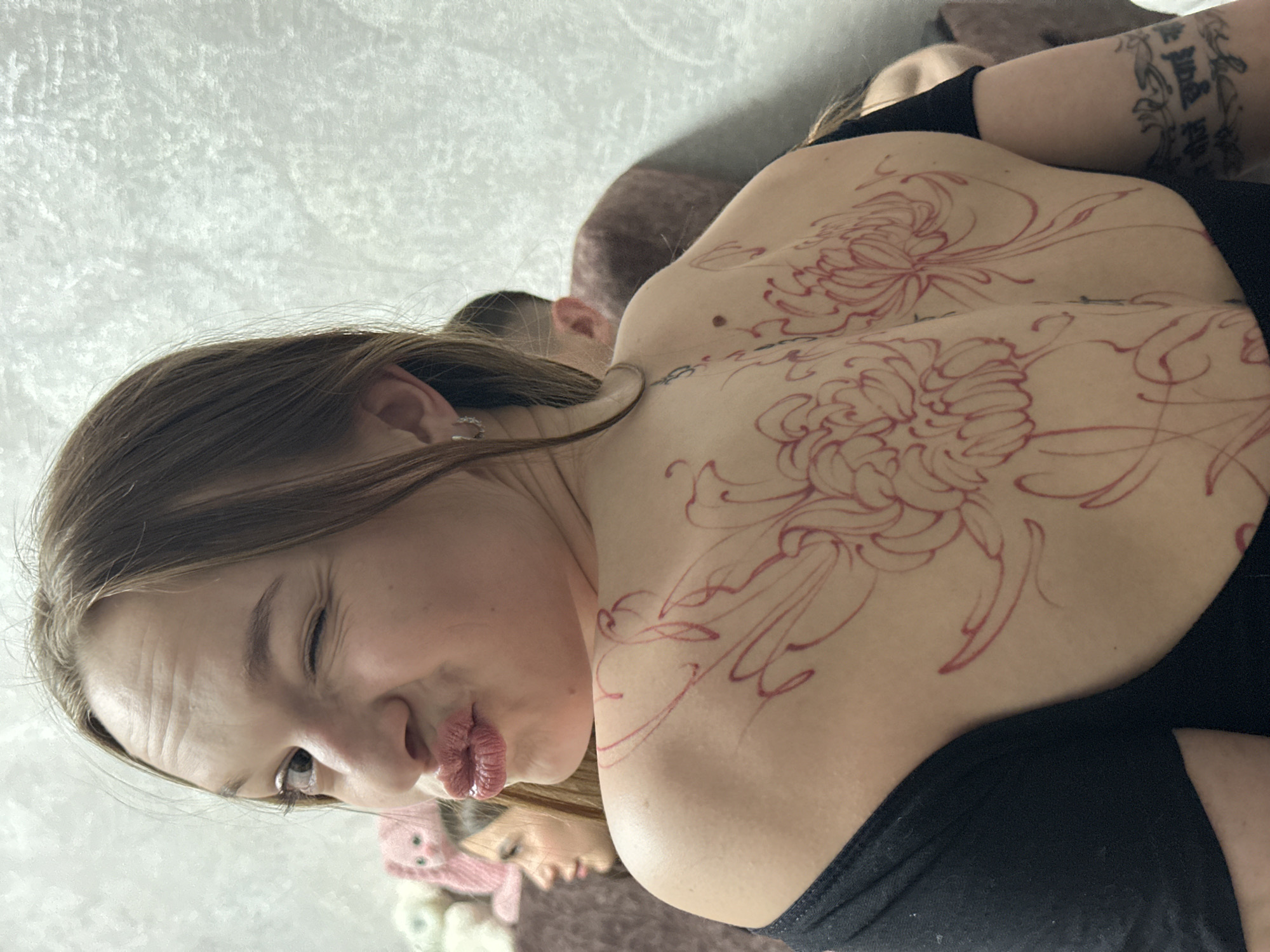 A young woman with long brown hair poses for a photo indoors in Chaikovsky, Russia, making a playful 'kiss' face towards the camera. She is seen from a slightly elevated angle, with her head tilted to the left and her body positioned horizontally. Her upper back and shoulders are prominently displayed, featuring a large, intricate red line art tattoo design depicting two chrysanthemum flowers and swirling organic patterns, suggesting the initial stages of a tattoo application. She wears a black top or bra that covers her shoulders. A small mole is visible on her left shoulder blade. On her right upper arm, a finished black tattoo with script, resembling Devanagari or a similar Indic script, is visible. In the background, out of focus, another young woman is partially visible, lying down, possibly wearing a pink knitted hat with ears. The setting appears to be a comfortable indoor space, with a textured light-colored wall or blanket in the background. The primary subject has a small, sparkling earring on her left ear.