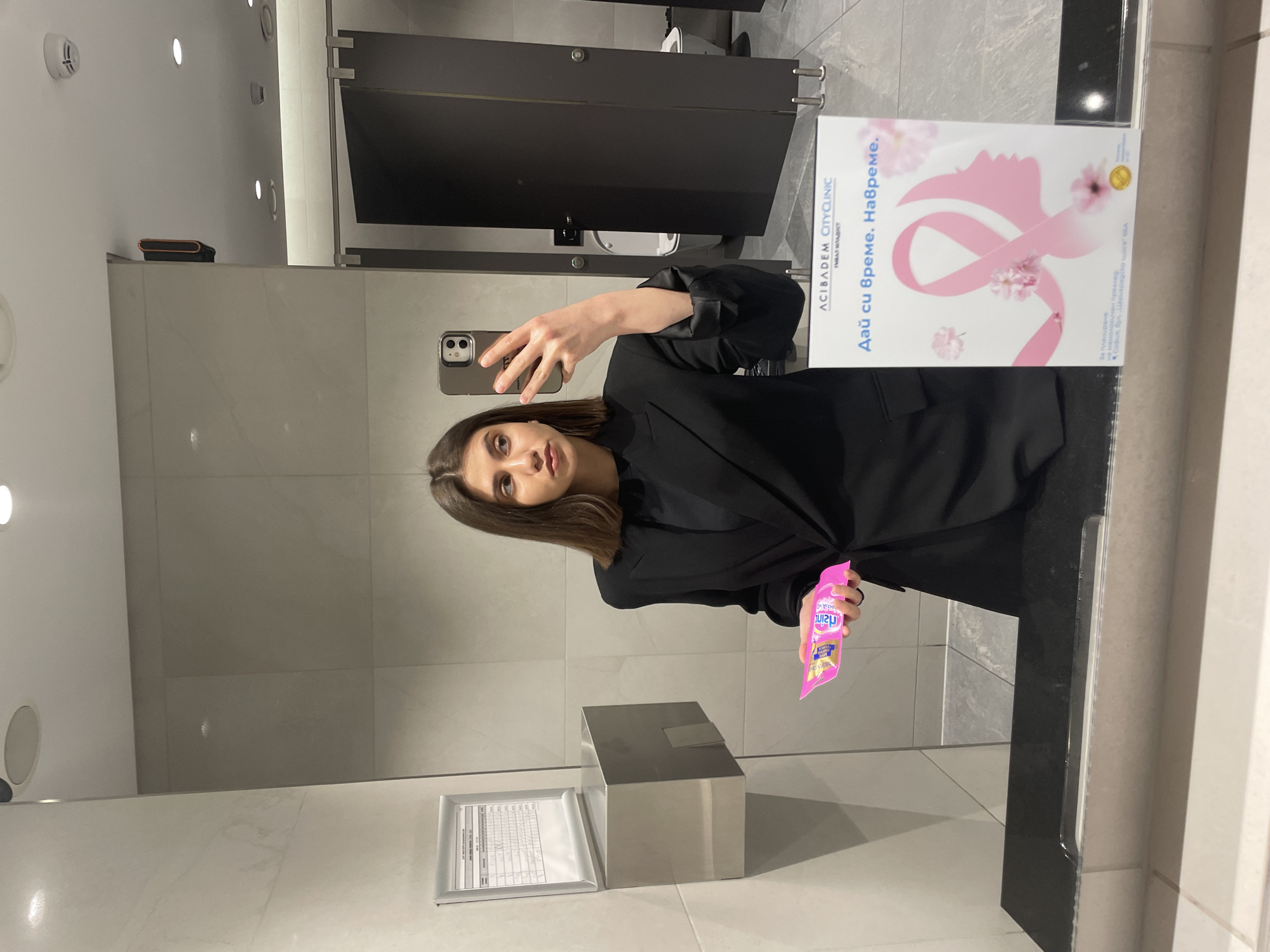 The image shows a woman taking a selfie in a restroom. She is holding a pink package of what appears to be candy or snacks. Behind her, a black bathroom stall door is visible. In the mirror, there is an advertisement for ACIBADEM CITYCLINIC УМВАЛ МЛАДОСТ. The sign says, "Дай си време. Навреме", which means "Give yourself time. On time". It notes the location of the clinic is "София, бул. „Цариградско шосе" 664", which translates to Sofia, "Tsarigradsko shose" Blvd. 664. The mirror reflects the restroom environment, including the tiled walls and various bathroom fixtures.