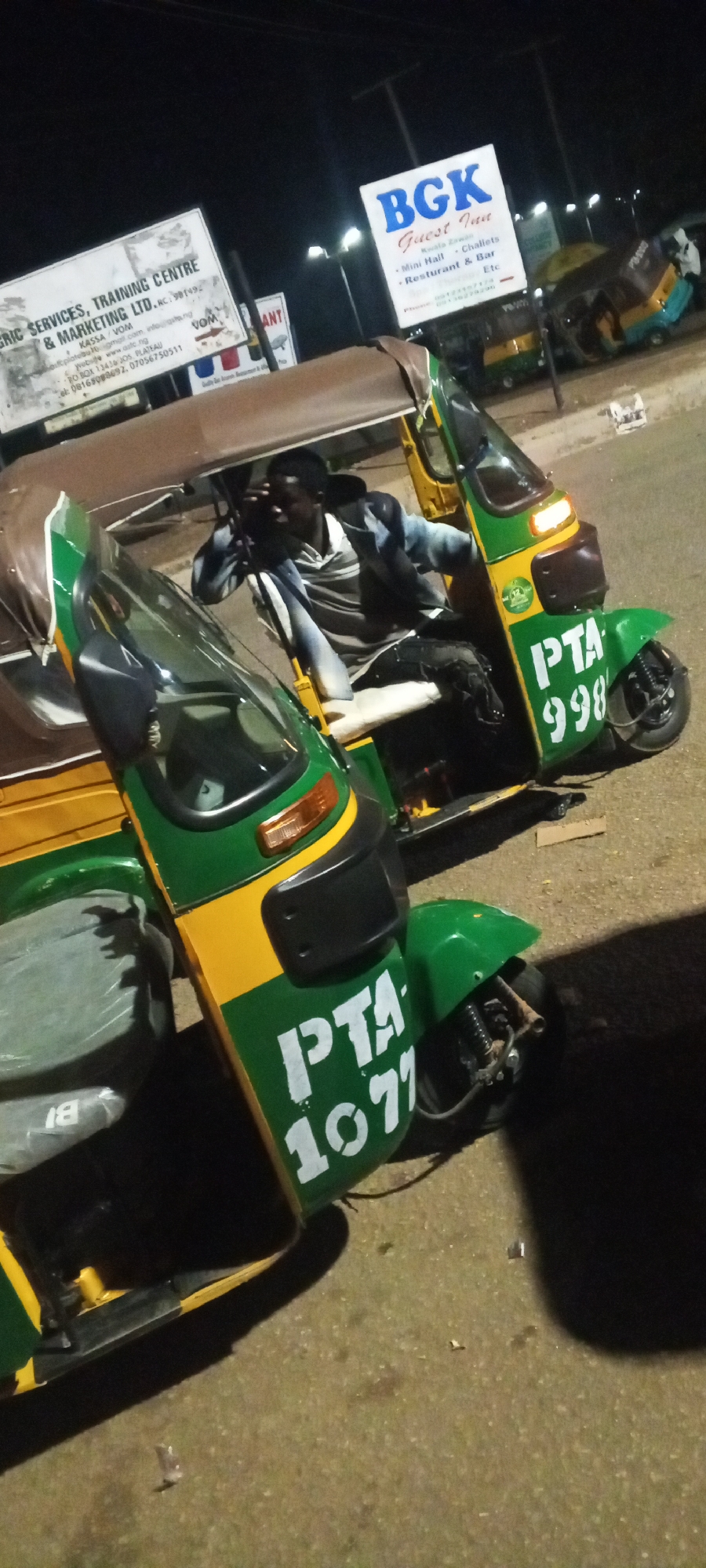 The image shows a nighttime street scene in an urban environment. Two auto rickshaws are prominently featured. The auto rickshaw in the foreground is green and yellow with "PTA 1077" painted in white on its side. A man wearing a striped shirt and a dark jacket is sitting in the driver's seat of this vehicle, looking away from the camera and shielding his eyes. The second auto rickshaw, partially visible behind the first, is also green and yellow and displays "PTA 998" on its side. In the background, there are several signs, including one that reads "BGK Guest Inn" and another with the text "ERIC SERVICES, TRAINING CENTRE & MARKETING LTD." The scene is illuminated by streetlights, casting shadows on the asphalt road surface. The overall impression is one of a busy street or parking area at night.