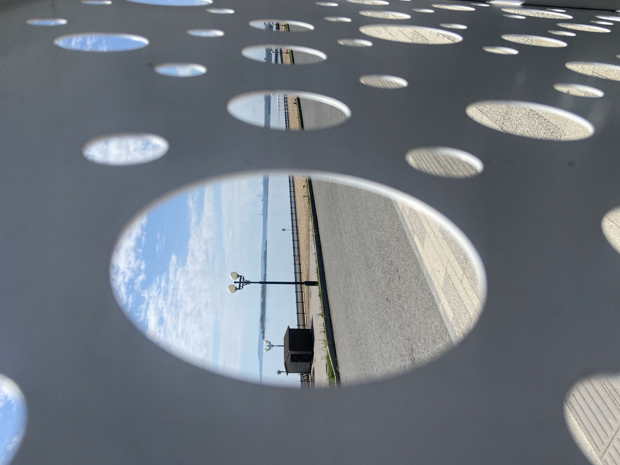 The image shows a view through a metal structure with various sized circular cutouts. The largest cutout frames a street scene in Saratov, Russia. The view includes a road, a walkway, a small structure or booth, a lamppost, a fence, and the Volga River in the background. The sky is visible with some clouds. The other cutouts show either the sky or portions of the walkway.
