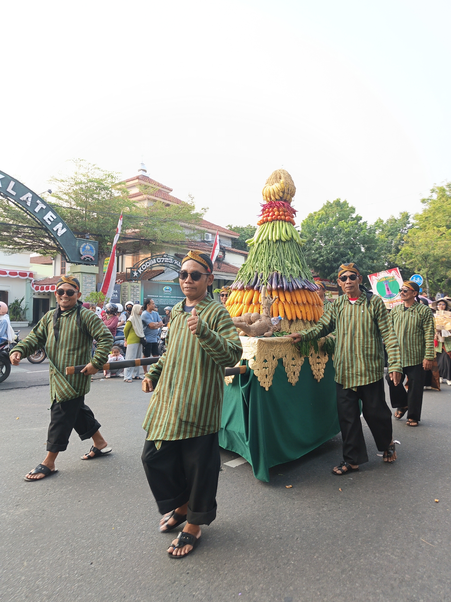 In Klaten, Indonesia, a parade is in progress. Four men wearing traditional Javanese outfits, consisting of striped long-sleeved shirts, black trousers, and headscarves, are walking on a street. They are wearing dark sunglasses and some are carrying a wooden pole. The men are escorting a large, cone-shaped float adorned with various fruits and vegetables, including corn, bananas, tomatoes, and what appear to be green beans and eggplants. The float is draped in green fabric with a gold trim. In the background, there are buildings, trees, and other people observing the parade. An archway with the text "KLATEN" is visible on the left side of the image. The sky is bright and appears to be daytime.