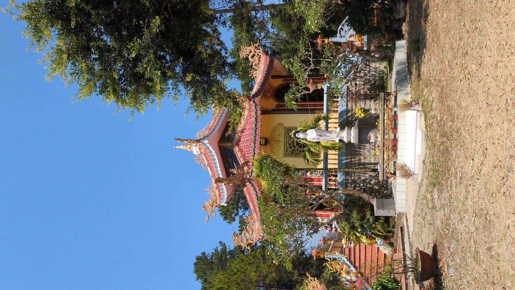 The image depicts a Buddhist temple in Lâm Sơn, Vietnam. The temple features a multi-tiered, ornate roof with decorative dragon motifs. A white statue of a deity stands in front of the temple. Vegetation surrounds the building, and a clear blue sky is visible above. The grounds appear dry and covered with foliage.