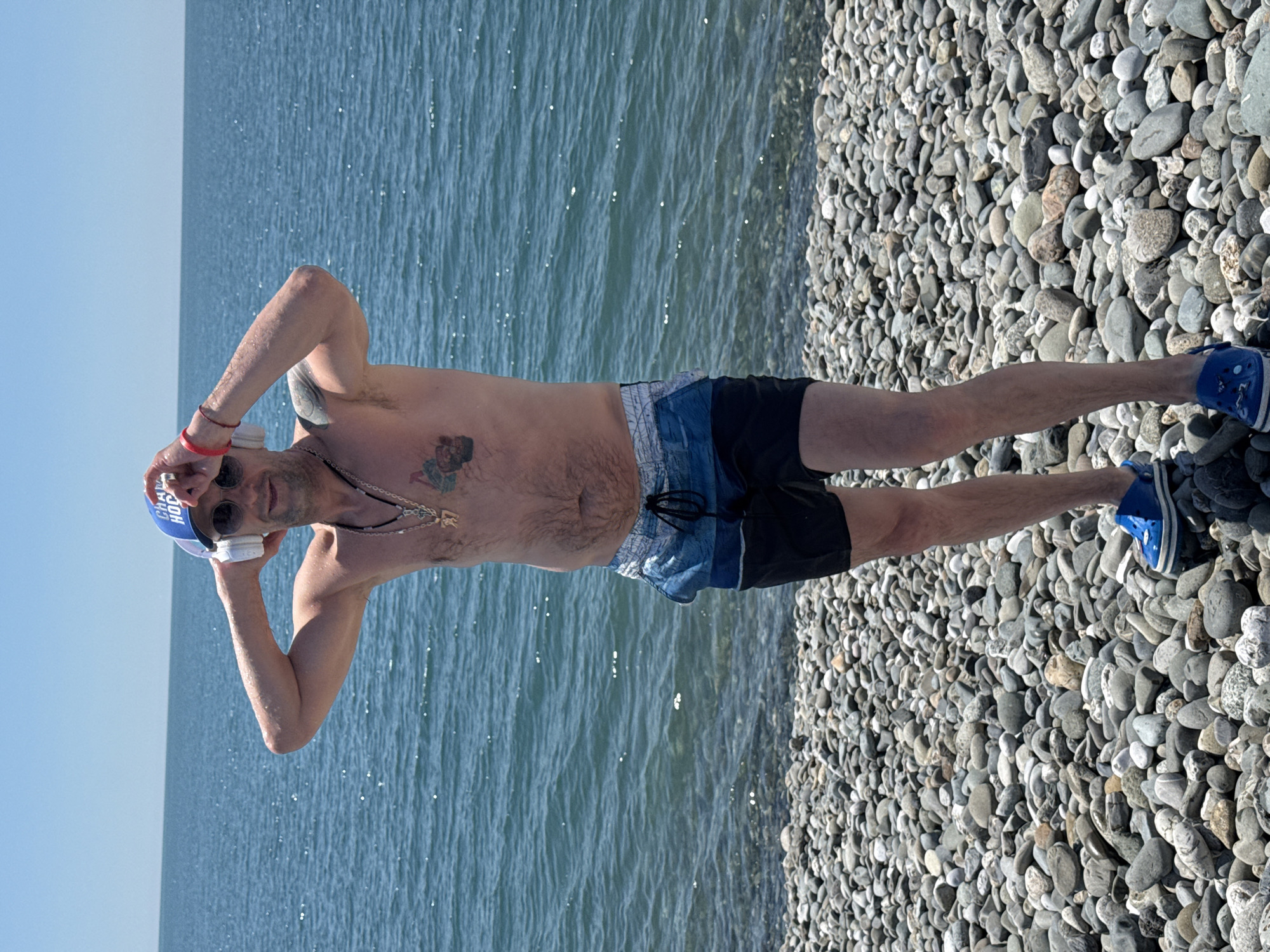 A man stands on a pebble beach by the sea, wearing shorts, a blue baseball cap, sunglasses, and blue Crocs. He has a necklace and a tattoo on his chest. He is holding white headphones on his head. The sea is calm with a bright sky overhead. The scene is likely near Sochi, Russia.