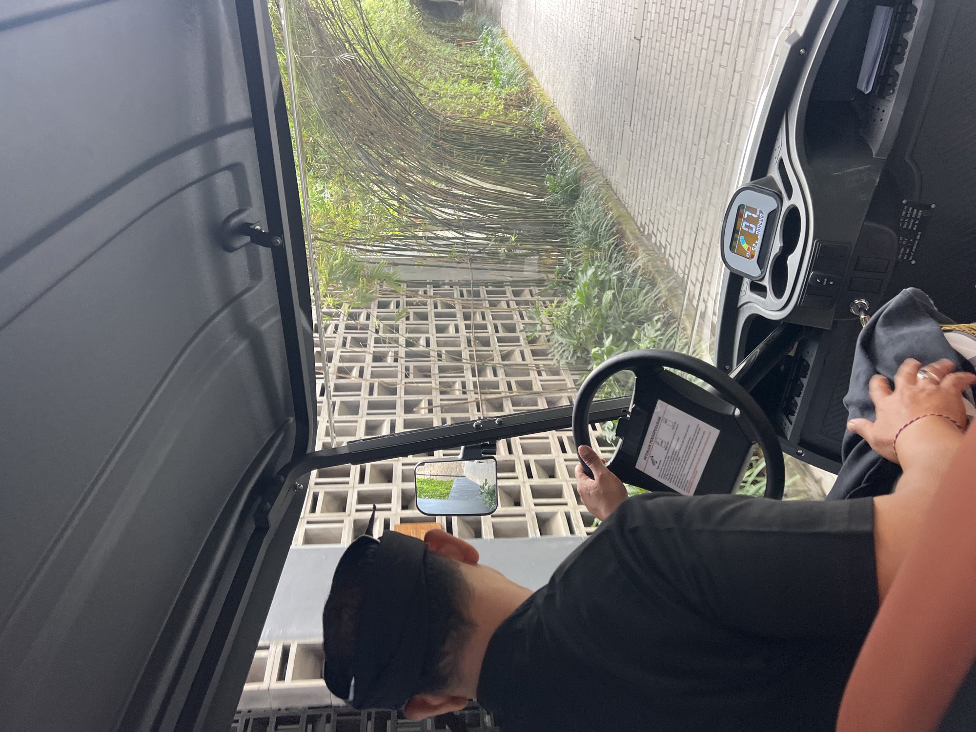 The image shows a man driving a golf cart along a paved pathway lined with bamboo trees and a patterned wall. The man is wearing a black uniform and a black head covering. He is holding the steering wheel and looking forward. The golf cart has a digital display that shows "07" and other information. The pathway appears to be in a tropical setting. This scene is likely located in Ubud District, Indonesia, given the distinctive architecture and lush vegetation. The overall impression is one of a leisurely ride through a serene and well-maintained environment.