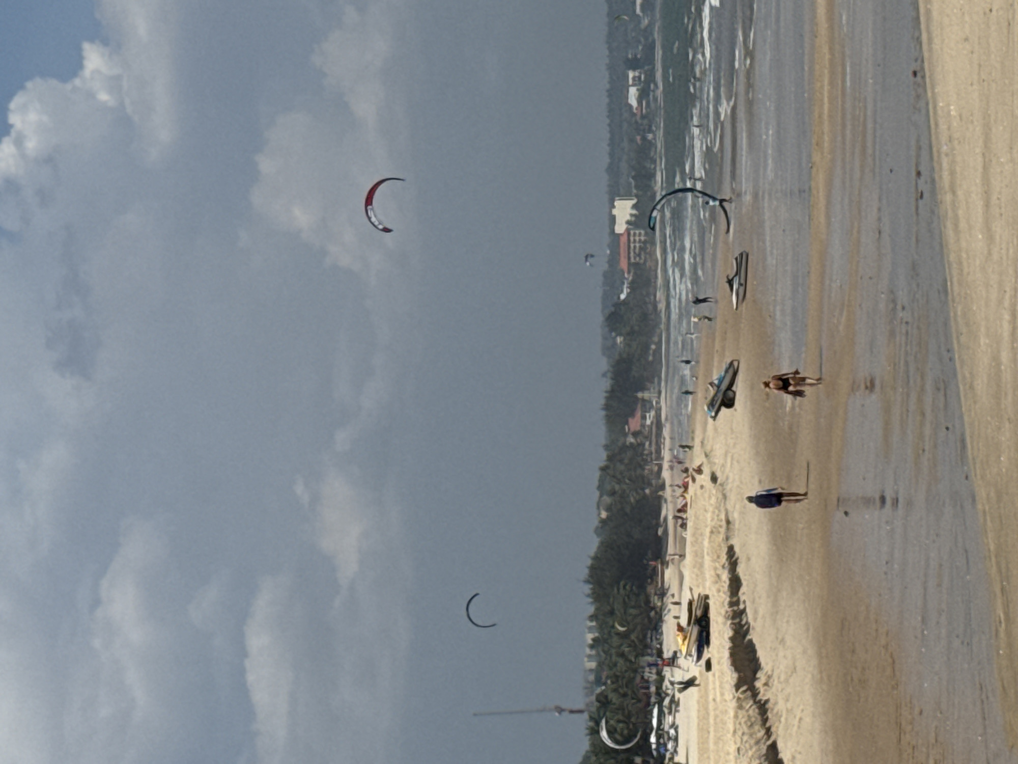 This is a wide-angle, eye-level shot of a beach in Phan Thiet, Vietnam, under a cloudy sky. In the foreground, a wet sandy area reflects the sky. Further up the beach, dry sand stretches out. Several people are visible walking along the beach. A woman in a dark swimsuit walks towards the viewer on the right, and a person in a dark blue and white outfit walks away from the viewer on the left. In the middle ground, several objects are present: a jet ski is parked on the sand, and what appears to be a surfboard or kiteboard is lying on the beach. Further down the beach, near the water's edge, a person is holding a kiteboard.
In the background, the ocean is visible with waves. Several kites are flying in the sky, indicating kiteboarding activity: a red and white kite is prominently displayed in the upper right quadrant, and a black kite is visible further to the left. Beyond the beach, a line of palm trees and buildings can be seen, typical of the coastal scenery in Phan Thiet. A tall structure, possibly a radio tower or a lookout, is also visible on the left side of the image behind the trees. The overall scene depicts a beach day with various recreational activities taking place.