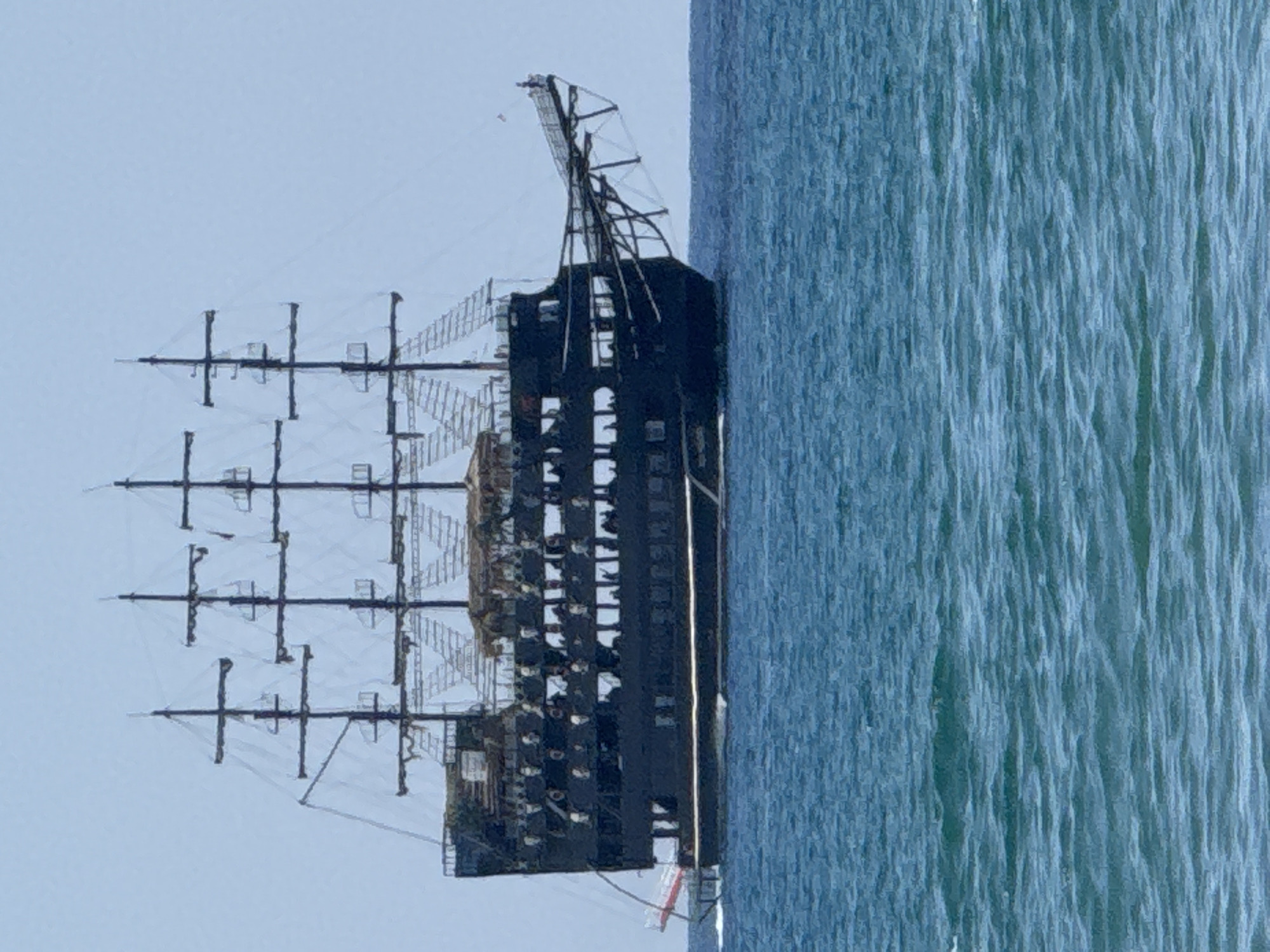 A large, black, multi-masted sailing ship is shown from its port side, moving through choppy blue water under a pale blue sky. The ship has several decks and appears to be a replica or themed vessel, possibly a pirate ship, given its dark hull and ornate rigging. A red and white gangway extends from the stern. The ship is likely at sea or in a harbor along the coast of Manavgat, Turkey, which is known for its tourism and coastline. The image captures a clear day with calm seas apart from the wake of the vessel.