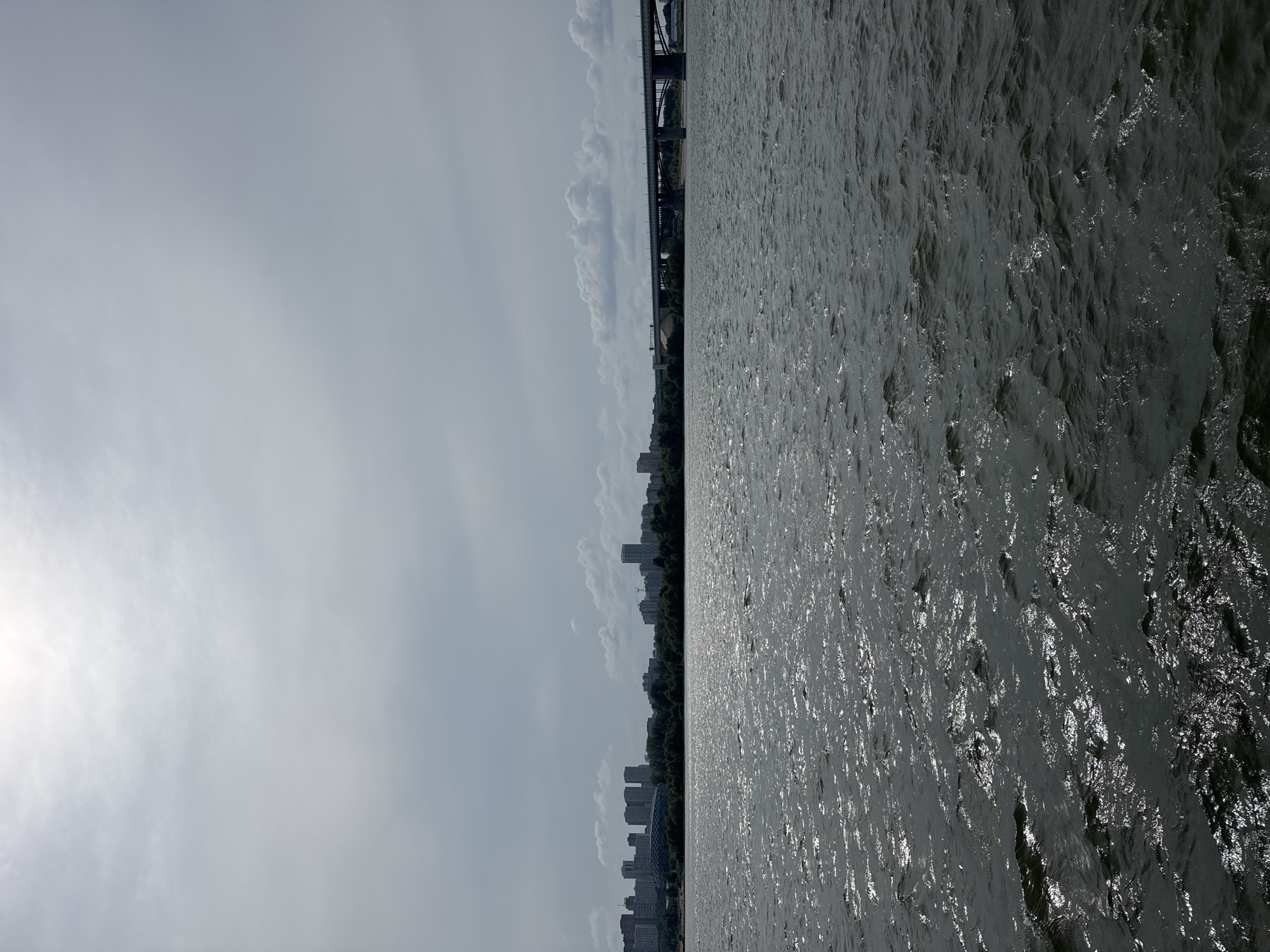 The image depicts a wide body of water, likely the Ob River, in Novosibirsk, Russia. The foreground is dominated by the dark, choppy water, reflecting the overcast sky and creating a textured, shimmering surface. In the mid-ground, a dense line of trees forms a dark band along the far bank. Beyond the trees, a city skyline is visible, characterized by numerous tall buildings and apartment complexes. To the right of the city, a long, multi-span bridge stretches across the river, with its structure clearly defined against the sky. The sky itself is a hazy expanse of gray clouds, with a brighter, diffused light source suggesting the sun is behind them. There are no people or animals visible in the scene. The overall impression is of a somewhat moody, urban landscape viewed from the water on a cloudy day.
