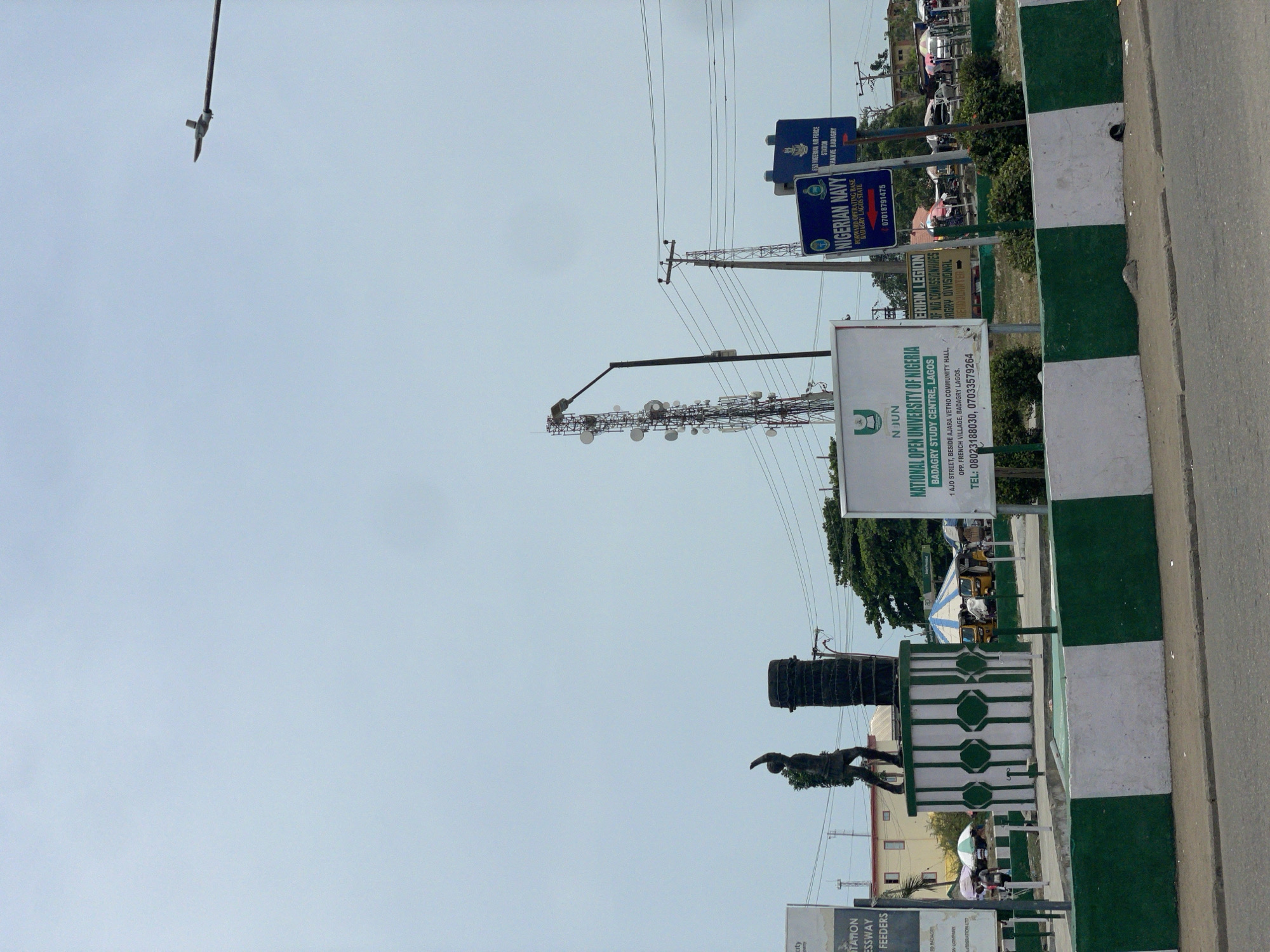 The image shows a street scene in Badagry, Nigeria. A green and white curb lines the road. Roadside signs are visible for the National Open University of Nigeria (NOUN), the Nigerian Legion, the Nigerian Navy, and the Nigerian Air Force. A statue sits on a green and white-painted base. A telecommunications tower is nearby.