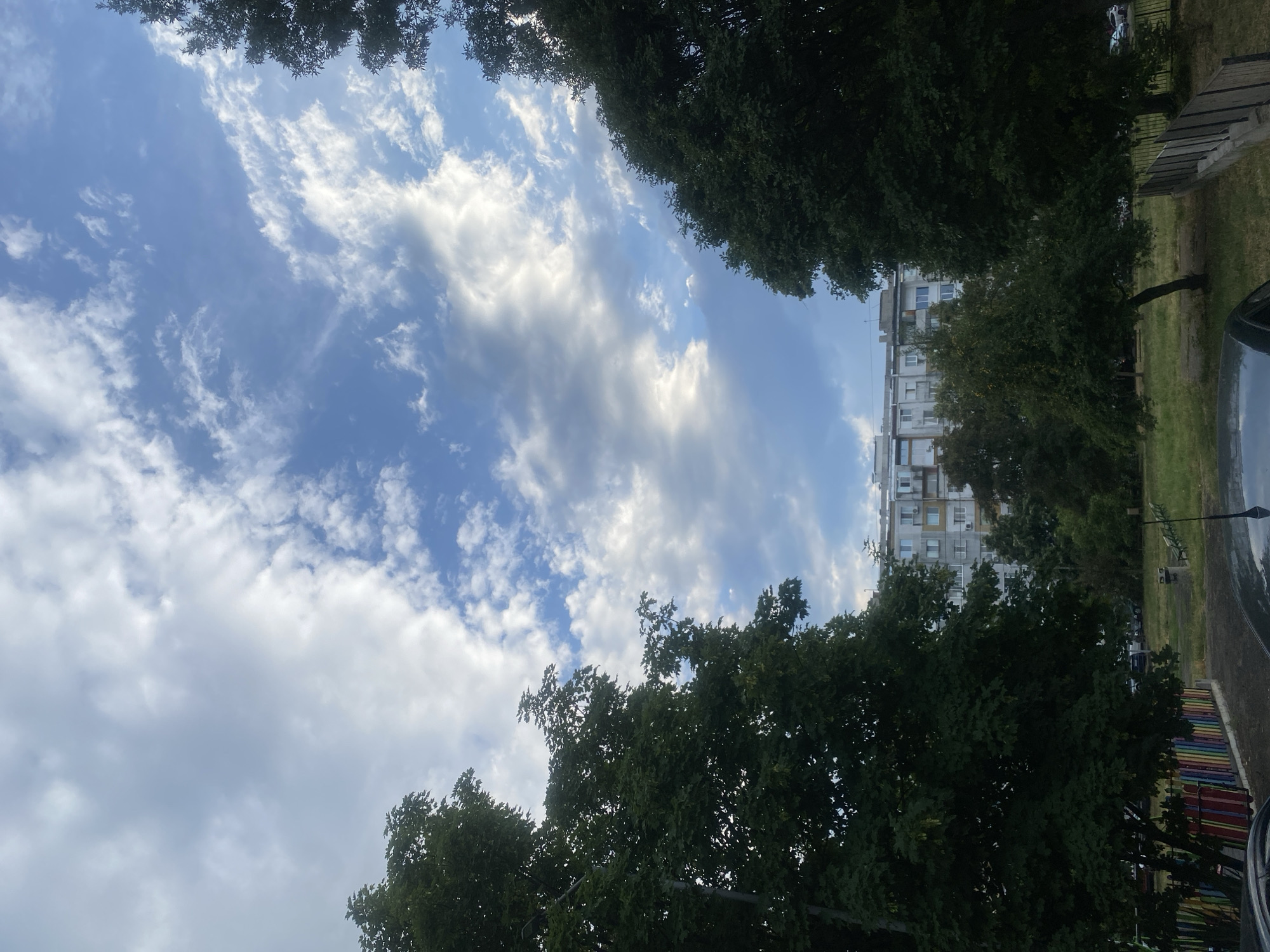 The image depicts a street view in Burgas, Bulgaria, under a partly cloudy sky. In the foreground, two large, dense trees frame the scene, one on the left and one on the right, with their dark green foliage obscuring a significant portion of the view. Between the trees, a multi-story apartment building is visible in the mid-ground, characterized by its light-colored facade and numerous windows, some of which appear to have air conditioning units. The building is set back from the street, with a grassy area and smaller trees in front of it. In the immediate foreground, the top of a dark-colored car and a portion of a colorful fence, possibly part of a playground, are visible. A few benches are present in the grassy area between the apartment building and the fence. The sky is a blend of blue and white clouds, with patches of sunlight breaking through, suggesting a dynamic weather condition. The overall impression is an urban residential area with mature trees contributing to the landscape.
