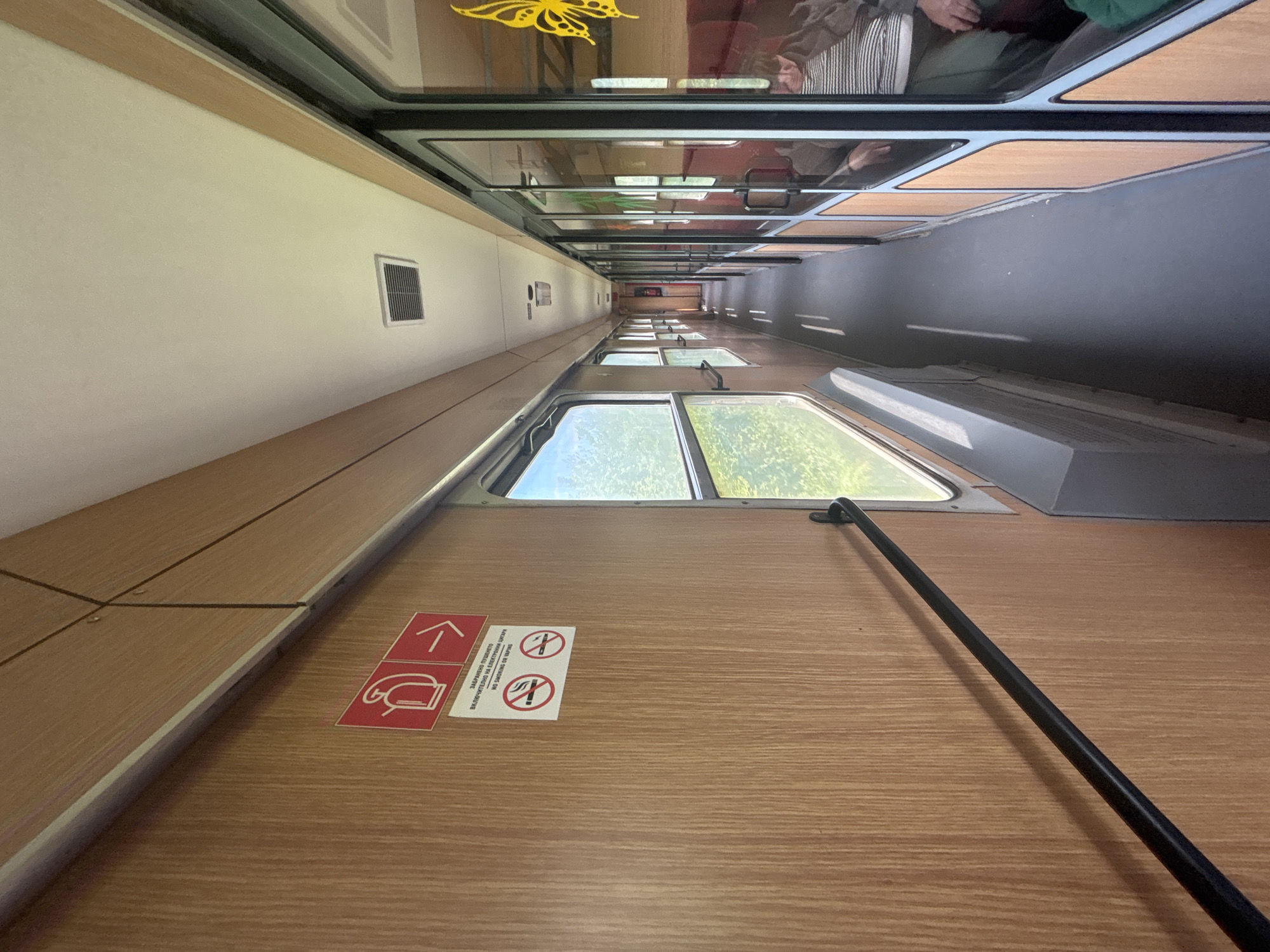 A view of the interior of a train car. The walls are paneled, and a window shows green trees outside. Overhead storage racks are visible, and people are seated in the racks above. Signage prohibiting smoking is posted on the wall. A metal bar runs diagonally across the foreground. The location is not discernible from the given image.