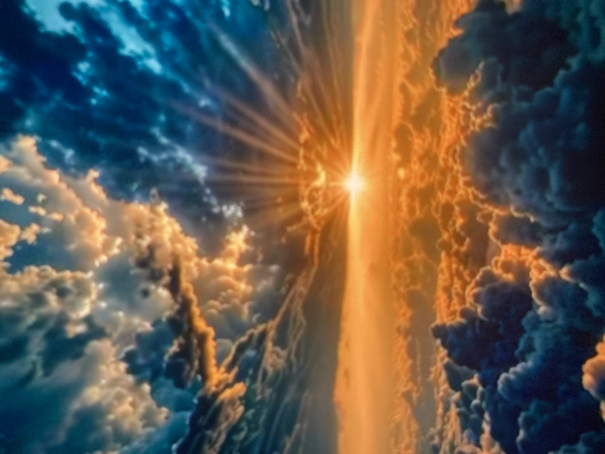 The image depicts a dramatic sky scene, likely captured from an aerial perspective. The dominant features are expansive clouds illuminated by a vibrant sunset or sunrise. Sunlight bursts forth from a central point, casting golden and orange hues across a sea of clouds. Some clouds are tinged with deep blues and grays, creating a striking contrast with the warm tones. The sky transitions from a brilliant orange horizon to darker blues and teals towards the upper edges of the frame. There are no discernible people, subjects, objects, or actions within the image. The scene is purely environmental, showcasing the dynamic beauty of atmospheric conditions above what would be the city of Ustyvytsia, Ukraine.