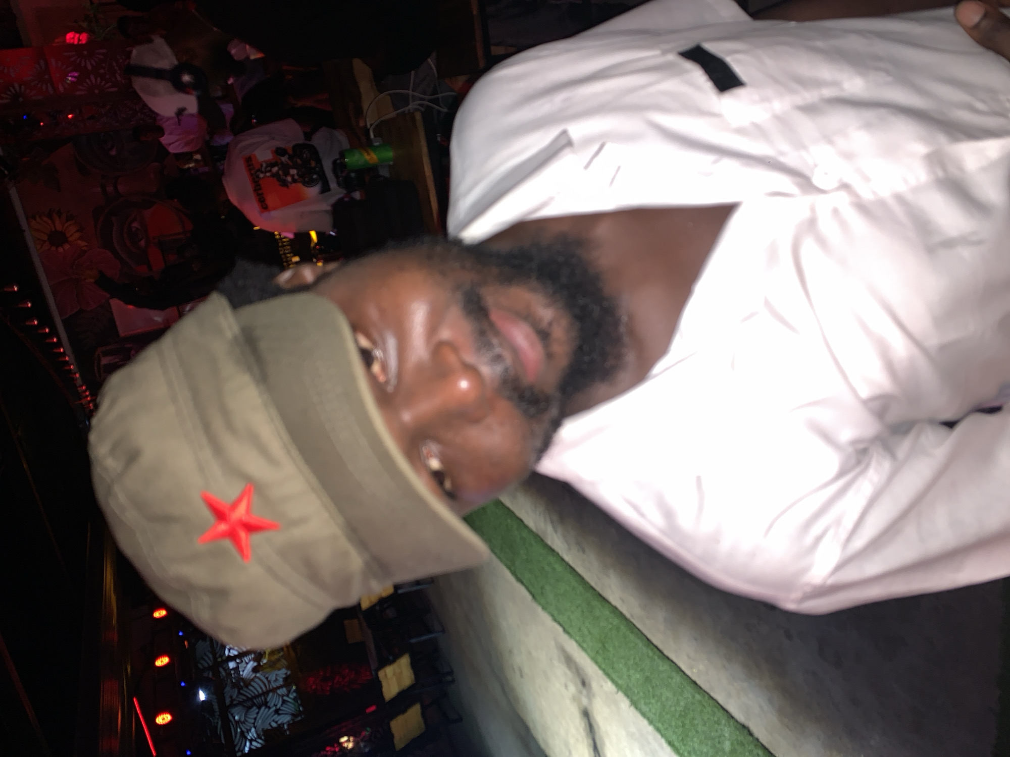 A close-up, slightly angled, head-and-shoulders outdoor, nighttime, portrait shows a man wearing a khaki cap with a red star emblem, and a white collared shirt. He has dark skin, a beard, and is looking towards the camera. The background is dimly lit and appears to be an outdoor seating area at night, possibly a bar or restaurant, with some tables and chairs visible, as well as a mural on a wall. There are strings of lights and some artificial turf visible in the scene.