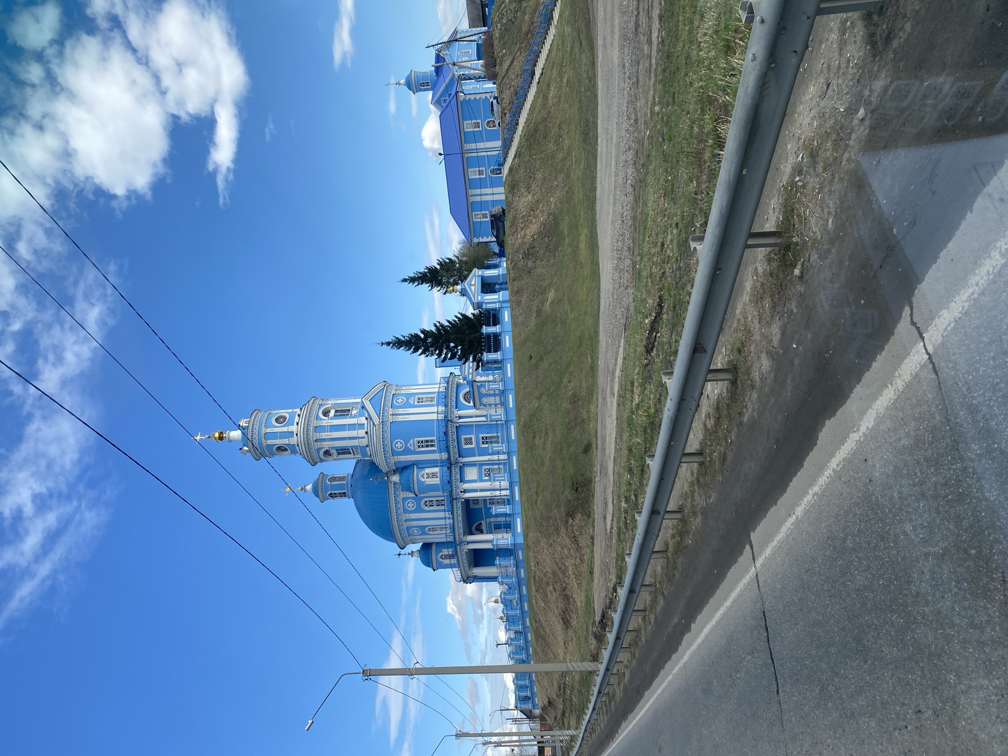The image shows a blue and white orthodox church in Telma, Russia, captured from a roadside perspective. A grey guardrail runs along the right edge of the frame, next to an asphalt road. Beyond the guardrail, there's a strip of green grass and dirt. The church stands out with its blue domes and white architectural details, contrasting against a clear blue sky dotted with scattered white clouds. Electrical power lines run diagonally across the sky in the lower left part of the image. A secondary, smaller blue building can be seen behind and to the right of the main church building. The overall scene is captured in daylight.