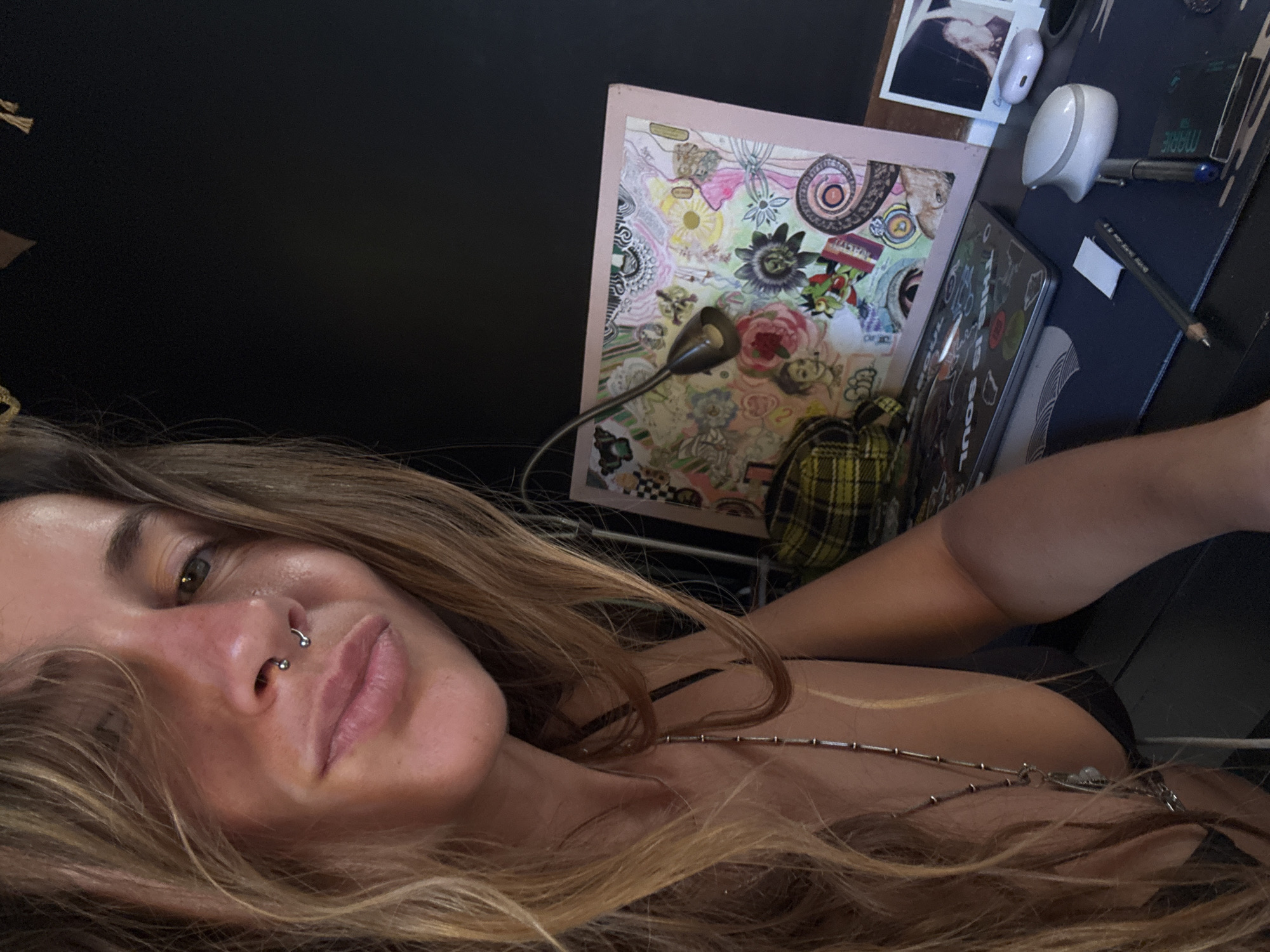 A woman with long blonde hair and a septum piercing is the main subject in this selfie, taken at an unknown location. She is looking towards the camera with a relaxed expression. Her hair is loose and falls around her face and shoulders. She is wearing a necklace with small beads and a pendant.
In the background, a desk is visible with various objects. A laptop with stickers on it is positioned on the desk. To the left of the laptop is a framed piece of art with colorful floral and abstract designs. A small, gooseneck desk lamp is attached to the frame. Further to the right, a white computer mouse and a pen are placed on a dark desk mat. A small photograph is also visible on the desk, near the top right corner of the frame. A plaid fabric, possibly a shirt or bag, is draped over part of the desk.
The lighting suggests it could be daytime or artificial lighting, as there are no strong shadows indicating natural sunlight. The overall mood is casual and personal. No text is readily identifiable in the image, except for some illegible text on stickers on the laptop.