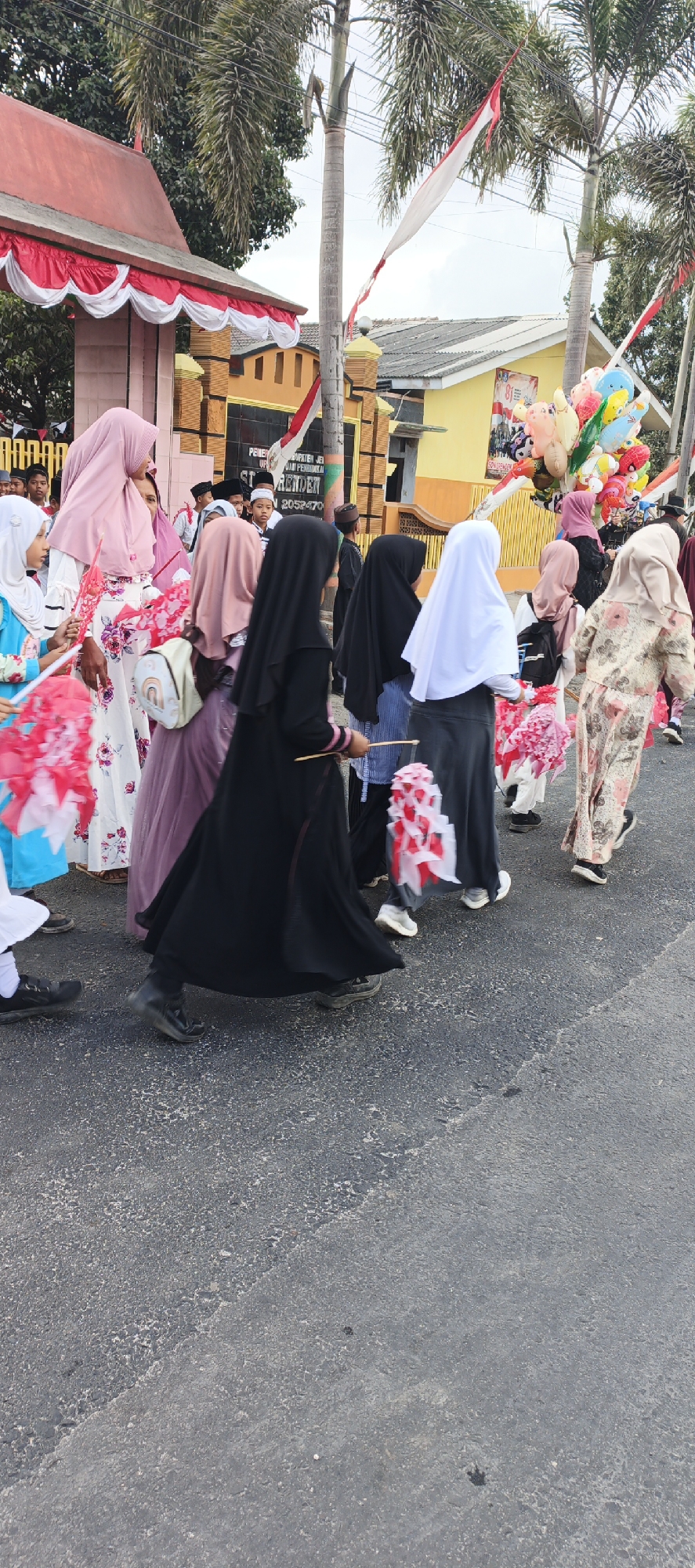 A parade is taking place in front of a building with red and white decorations. The building appears to be a government or educational institution, with signage visible that includes "PEMERINTAH KABUPATEN JEMBER" and "ST. HENDRA" along with a phone number. Several people are walking in procession, many of whom are women and children wearing Islamic attire, including hijabs. Some are carrying red and white decorative items, possibly flags or pinwheels, and a bunch of colorful balloons are visible in the background. The event seems to be festive, with patriotic decorations. The ground is paved asphalt. The location is identified as Grenden, Indonesia.