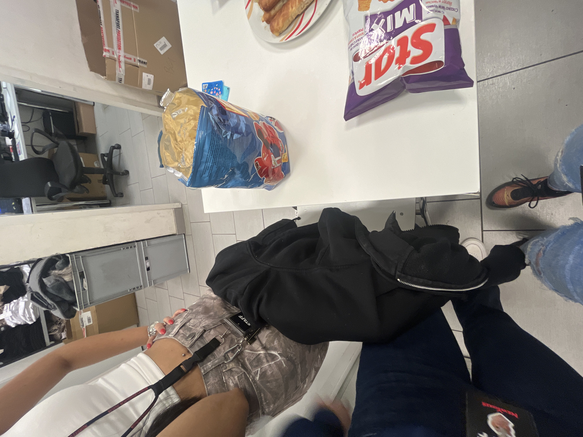 The image shows an overhead view of a table setting with two individuals present.
**Environment:** The scene appears to be indoors, likely a break room or casual dining area, identifiable through the tile flooring and the presence of office-like furnishings in the background.
**Objects:** On the white table, there's an opened bag of blue chips, a bag of Star Mix chips, and a plate with pastries. A black hoodie sits on the table near the chips.
**People:** Two individuals are partially visible. One person is wearing a white top and camo-patterned bottoms, with a "STAFF" tag visible. The second individual is wearing jeans and brown sneakers.
**Actions/Interactions:** The photo appears to be taken during a snack or break period, with the presence of chips and pastries suggesting eating or sharing food. No clear interaction is observable other than the shared space.
**Location (Contextual):** Although the location cannot be identified by just the photo, the chip brand is "Star Mix" and the chip name is labelled "Сладка Чили Чушка Йогурт и ЧестЫН", which suggests that the scene may be set in Bulgaria.