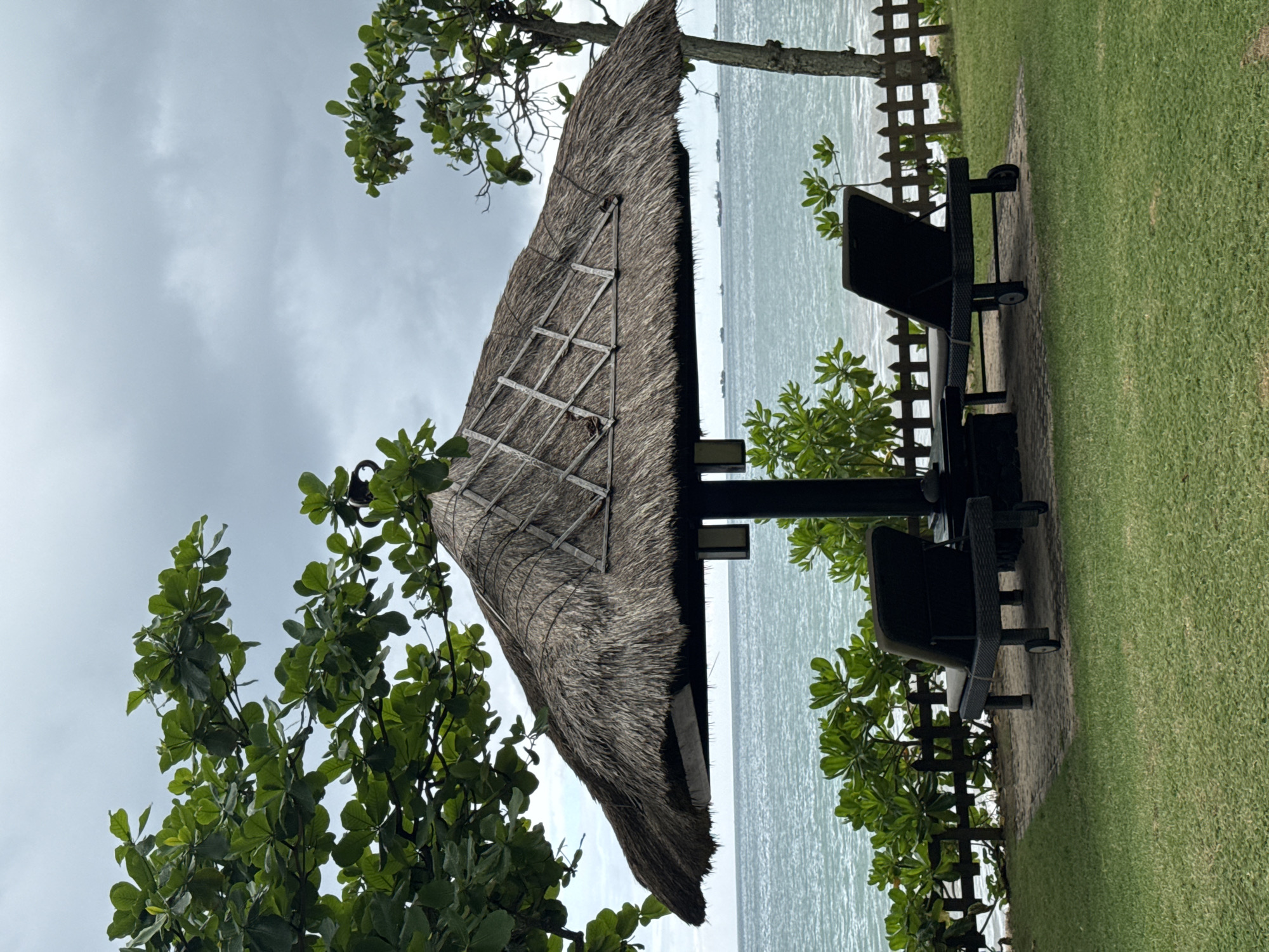 The image depicts a serene tropical or beachside setting in Kuta Selatan, Indonesia. The scene is dominated by a thatched-roof structure, possibly a gazebo or a part of a larger resort, positioned on a well-maintained patch of green grass. Two dark-colored lounge chairs are placed beneath the shade of this structure, facing the ocean.
In the foreground and midground, lush green foliage from trees frames the view, with some branches extending into the upper parts of the frame. A simple wooden fence runs along the edge of the grassy area, separating it from the expansive body of water beyond. The water appears calm to slightly choppy, reflecting the overcast sky. The sky itself is a soft grey with visible cloud cover, indicating a cloudy day, likely during daylight hours but without direct harsh sunlight.
No people are present in the image, contributing to a sense of tranquility and an inviting, undisturbed atmosphere. The arrangement of the chairs and the presence of the shaded structure suggest a peaceful spot for relaxation and enjoying the ocean view. No discernible text is visible within the image.