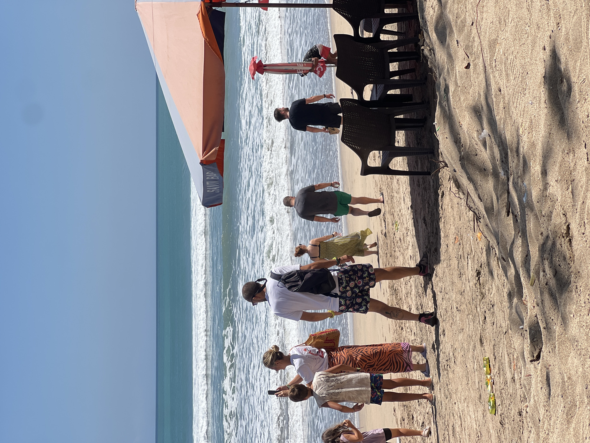 In Kuta, Indonesia, a sandy beach scene unfolds under a clear blue sky. The ocean, with its white-capped waves, extends to the horizon, meeting the sky. In the foreground, a beach umbrella with orange and white panels provides shade over several dark plastic chairs. A group of people are scattered across the sand. In the middle ground, a woman in an orange and black patterned skirt holds up a smartphone, seemingly taking a photo, while a boy in a light-colored t-shirt stands beside her. A man with a backpack walks away from the camera, facing the ocean. Further down the beach, other individuals are visible, some walking, others standing. Small, rectangular offerings, likely made of green material with colorful adornments, are placed on the sand in the lower left portion of the image. Debris, including what appears to be a cigarette butt, is also present on the sand. The overall atmosphere suggests a sunny day at a popular tourist destination.