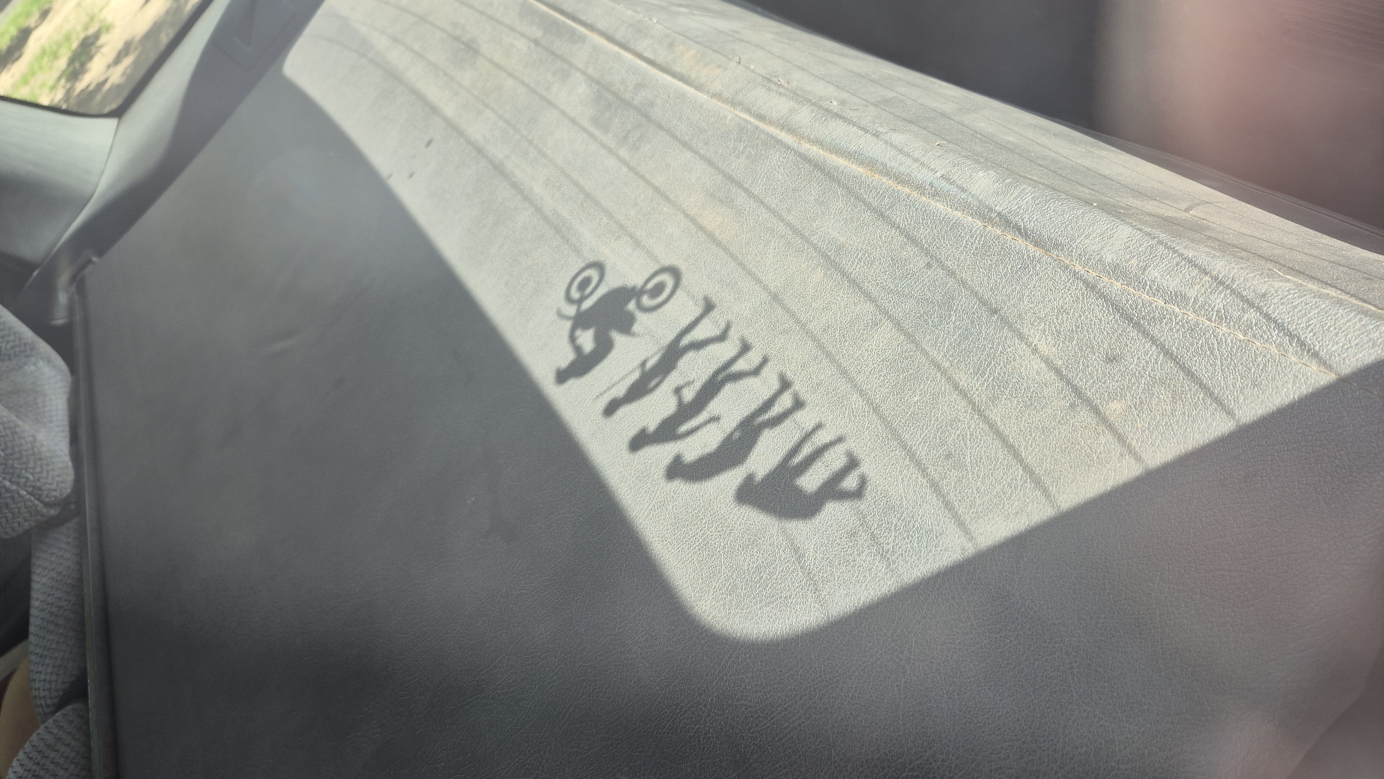 The image shows the shadow of the evolutionary progression of humans, from an ape-like figure to a person riding a dirt bike, cast across the sunlit rear seat of a vehicle. The seat is upholstered in a dark, textured material and has horizontal ribbed lines. The shadows are projected onto the seat from the right side of the frame, with a diagonal beam of sunlight cutting across the lower portion of the image. The background is out of focus, showing hints of the interior of the car and a blurred outdoor environment. This scene is likely captured within the city of Homyel, Belarus, given the provided context.