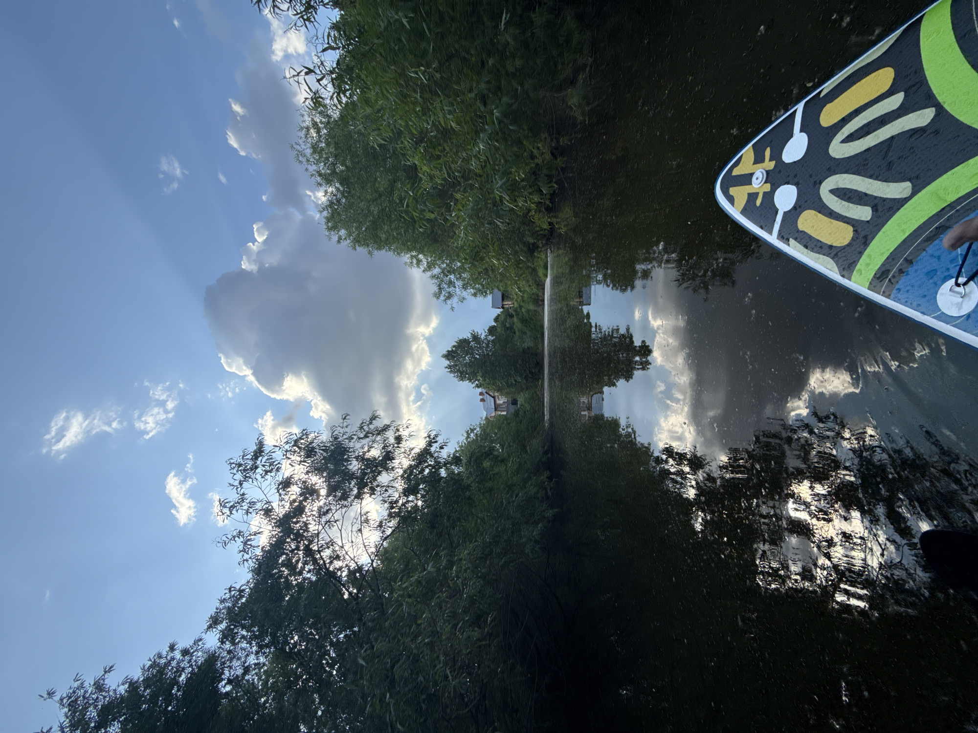 The image captures a serene view from a paddleboard on a calm body of water, likely a river or lake, near the village of Kondratovo, Russia. The water's surface reflects the sky and surrounding lush green trees, creating a mirror-like effect. In the distance, a red-roofed building is visible through a break in the trees. The sky is partly cloudy, with bright sunlight breaking through in some areas, casting rays that illuminate the clouds. The front of the paddleboard, adorned with a colorful abstract pattern, is visible in the foreground. The overall atmosphere is tranquil and natural.