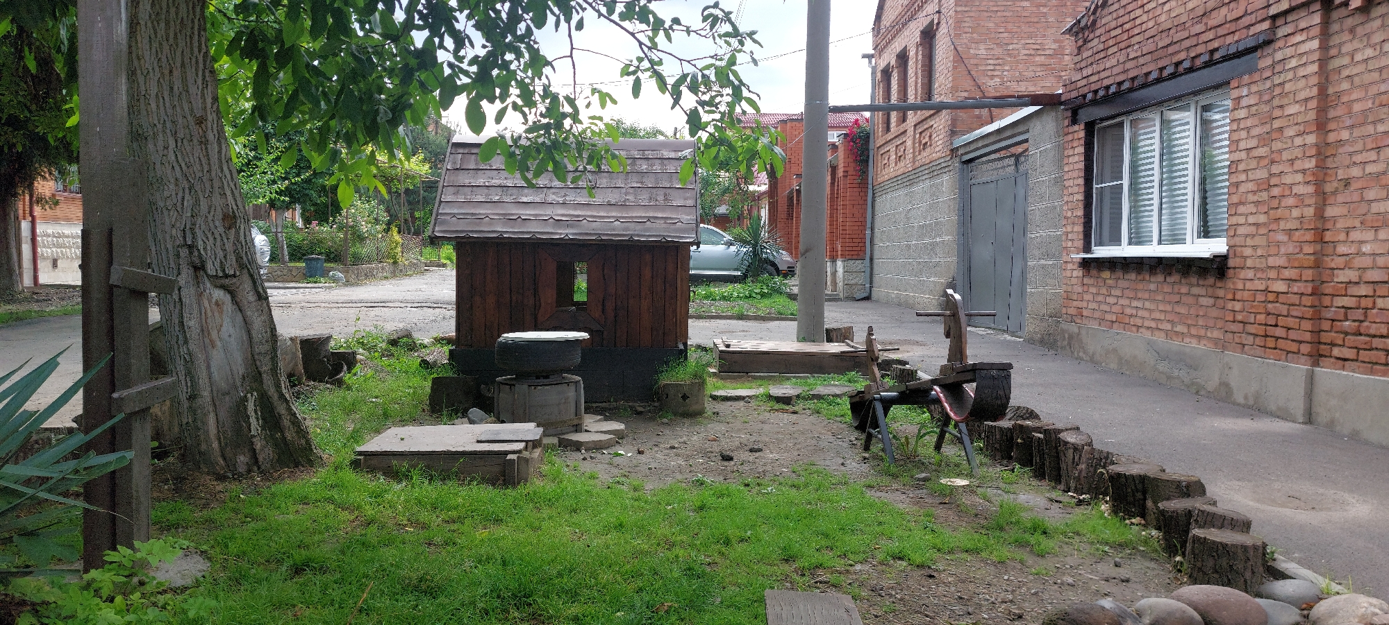 The image depicts an outdoor scene, likely a backyard or a small communal area, with a rustic and somewhat unkempt appearance. The central focus is a small, wooden, shed-like structure with a weathered, sloped roof, resembling a miniature house or a well cover. In front of this structure, there's a concrete wellhead topped with a tire and a metal lid.
To the left, a large tree with rough bark dominates the foreground, its branches and green leaves arching over the scene. A concrete pole stands next to the tree, appearing to be a utility pole. Further left, a paved path or road is visible, with a hint of other buildings and greenery in the background.
To the right of the wooden structure, a wooden seesaw-like toy, possibly homemade, is positioned. It appears weathered and has a red seat. A grey metal garage door is also visible on a brick building in the background. The ground is a mix of grass and dirt, with scattered pieces of wood, stones, and what look like tree stumps lining a pathway.
The lighting suggests it might be daytime, possibly overcast, as there are no strong shadows. The overall atmosphere is quiet and appears to be a residential or a rural setting. No people are visible in the image. There is no visible text.