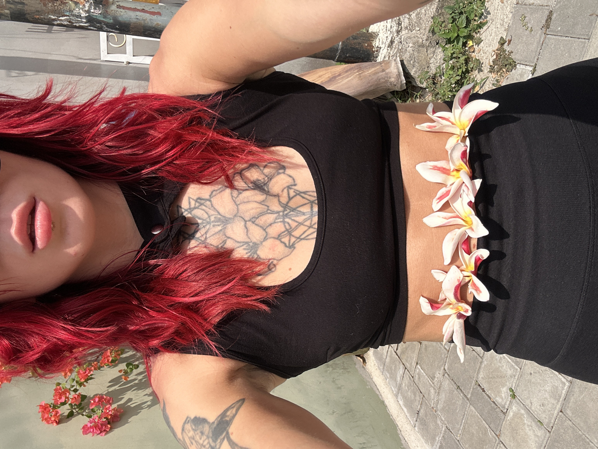 A person with vibrant red hair, wearing a black crop top and black leggings, poses outdoors in North Kuta, Indonesia. The person has tattoos on their chest and left arm. Several plumeria flowers are arranged around their waist. The background features a textured wall, a paved walkway, and some greenery. The scene is illuminated by bright sunlight, casting shadows on the ground.