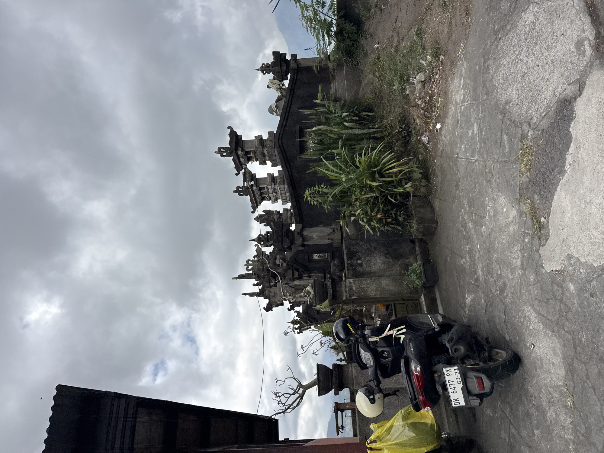 The image depicts a gray, overcast day in Kintamani, Indonesia. In the foreground, a black scooter with license plate DK 6477 PX is parked on a concrete surface. Two helmets, one white and one black, are attached to the handlebars. A yellow plastic bag is visible to the left of the scooter. Behind the scooter, a stone structure with intricate carvings and multiple tiers dominates the middle ground. Green plants, including snake plants, grow around the base of the structure. In the background, distant mountains are shrouded in mist. The overall scene suggests a cultural or religious site, possibly a temple complex, given the architectural style of the stone structure. The environment appears somewhat unkempt, with some debris visible on the ground.
