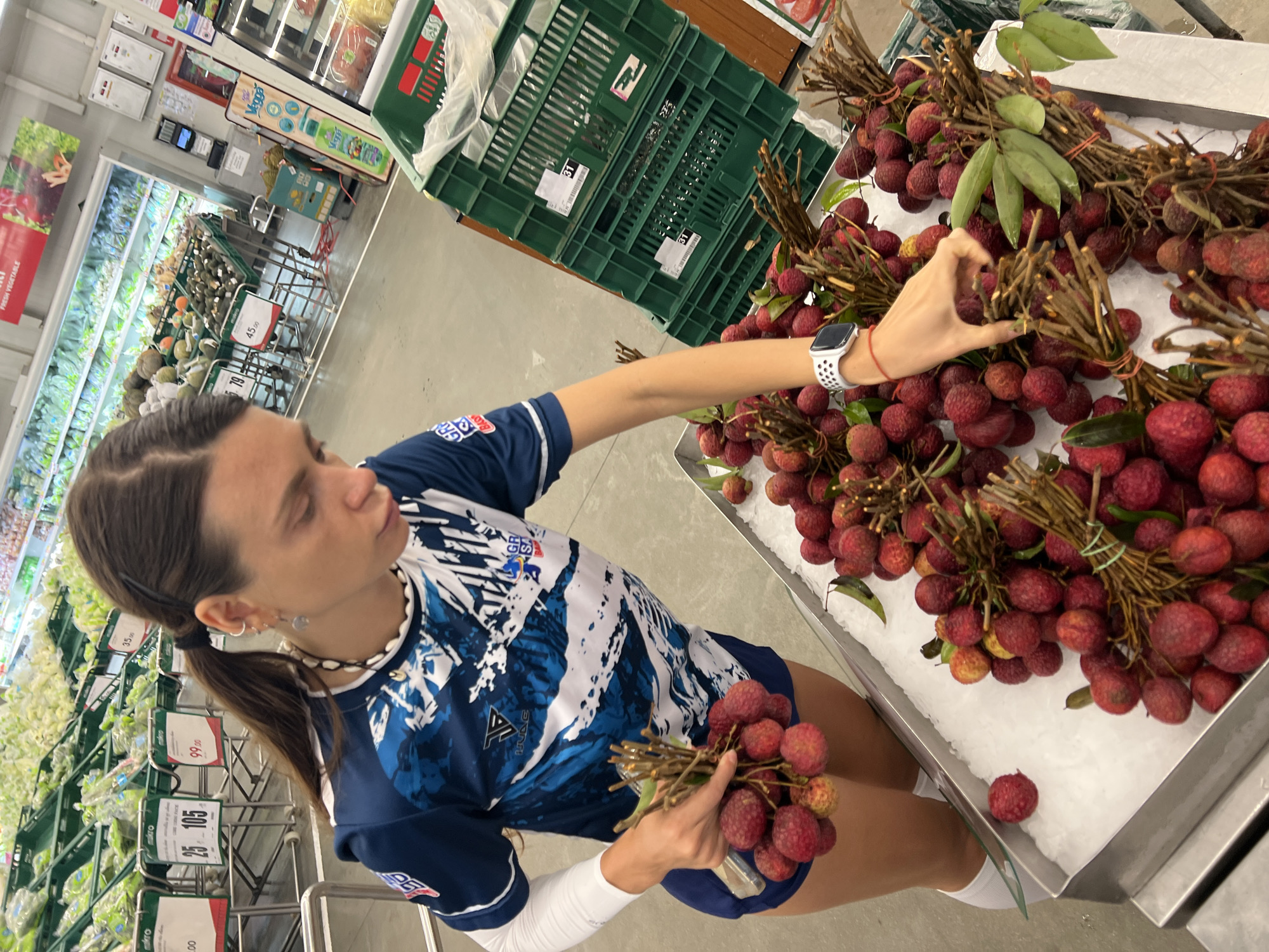 In Baan Bo Phut, Thailand, a person with dark hair tied in a ponytail and wearing a blue and white athletic shirt is in a supermarket. They are reaching for bunches of red lychees displayed on a bed of ice. The person is wearing a white smartwatch and a necklace. The background shows shelves stocked with fresh produce and green crates labeled with numbers. Several price tags are visible, including "25 105" and "99.00", and "45.00". The lighting suggests it is daytime. The overall scene depicts a shopper selecting fruit in a grocery store.