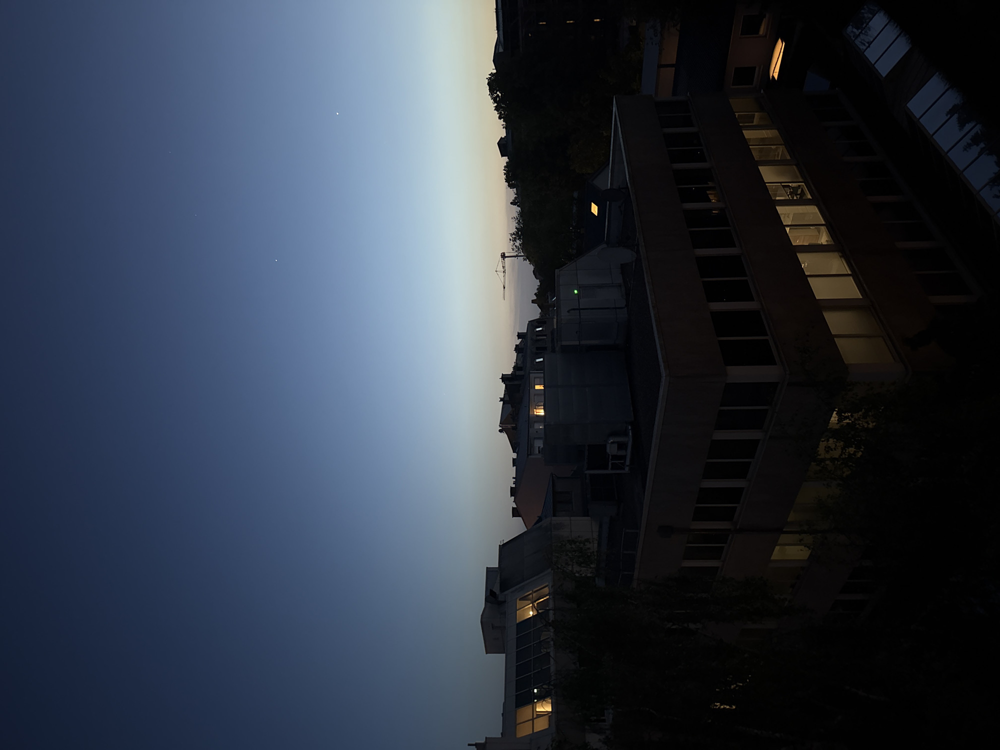 The image depicts a cityscape in Luxembourg, likely in the city of Luxembourg, at dusk or early evening. The sky transitions from a deep blue to a light gray near the horizon. Several buildings are visible, some with lights on in the windows, indicating occupancy. A construction crane can be seen on the horizon, suggesting ongoing development. The buildings vary in height and design, and there is some greenery visible, including trees. The scene is generally urban, with architectural features and infrastructure typical of a city environment.