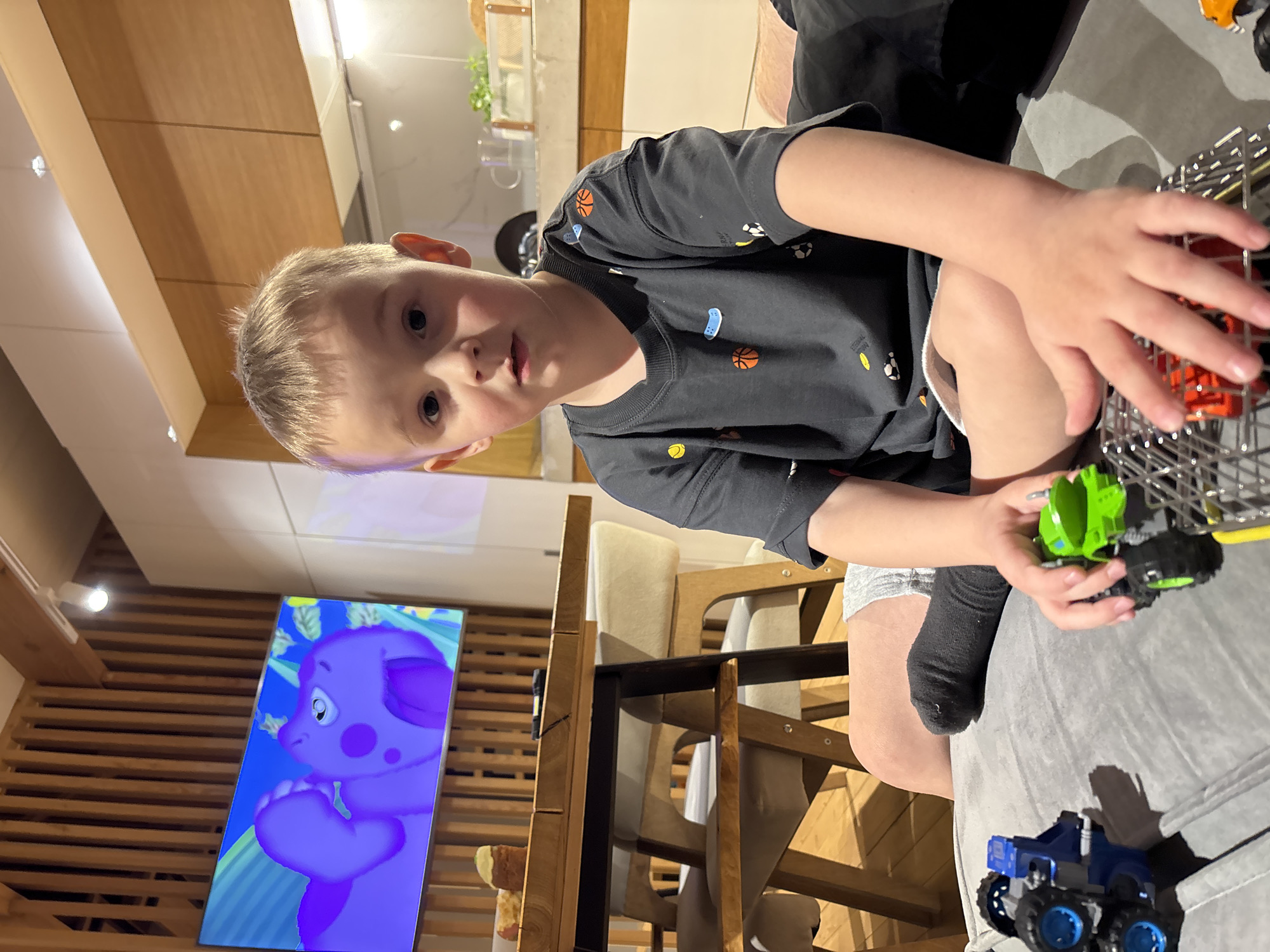A boy with blond hair is sitting and playing with toy cars. He is wearing a dark t-shirt with sports-themed graphics and grey shorts. He has one sock on. There is a cage-like container filled with toy cars. A green toy car is in his hand and a blue toy car is on a grey surface beside him. Behind him is a television playing a cartoon with a purple elephant-like character. The background includes a wooden wall, chairs, and a kitchen area with light-colored cabinets. The location is in a room with a modern interior. There is no information to suggest this scene occurred in the city of Chaikovsky, Russia.