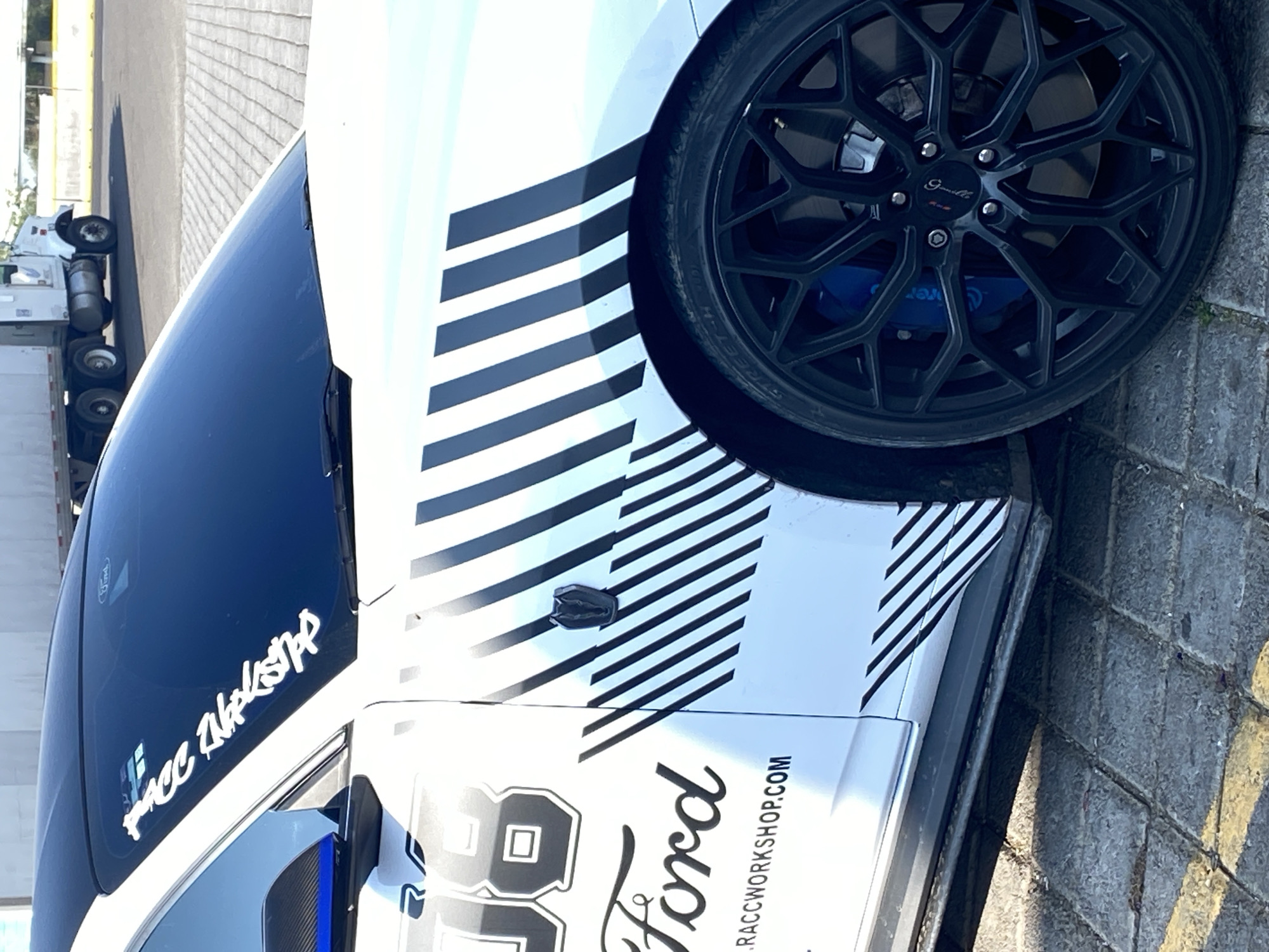 The image shows a partial view of a white Ford Mustang with black racing stripes on the side, parked on a paved surface. The car features a black rim with blue calipers and the number "86" with the word "Ford" prominently displayed, along with the website "RACCWORKSHOP.COM." A sticker on the windshield reads "RACC WORKSHOP." In the background, a truck is partially visible. The photo was taken in the city of Santa Ana Xalmimilulco, Mexico.