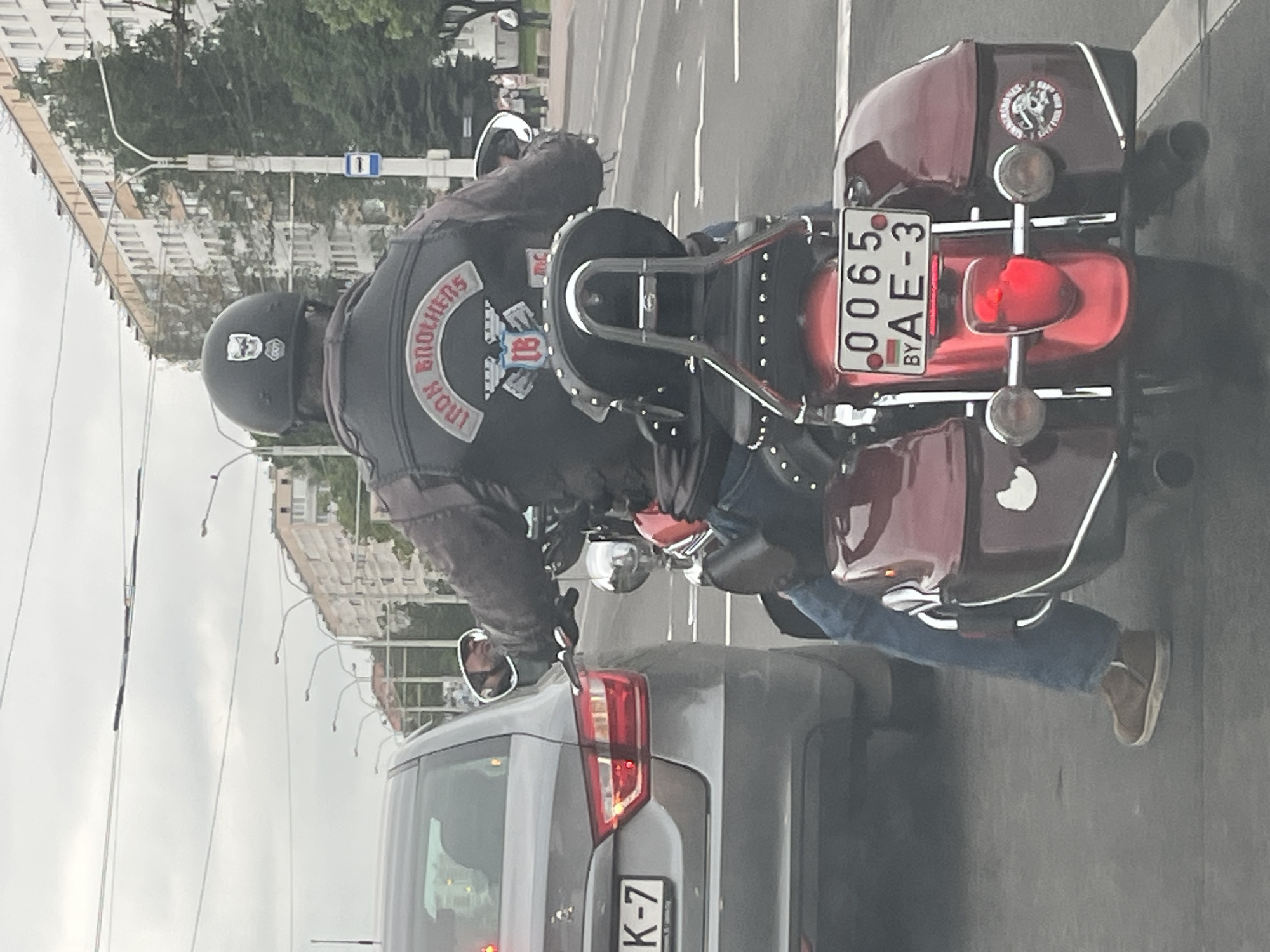 An overhead, slightly angled view captures a motorcyclist on a burgundy cruiser motorcycle on an urban street during daylight hours under an overcast sky. The motorcyclist, seen from the rear, wears a black leather jacket adorned with prominent patches, blue jeans, and brown shoes. A black helmet, featuring a white skull decal, covers the rider's head.
The most visible patch on the leather jacket is a top rocker that reads "IRON BROTHERS" in white text on a red background. Below it is a central emblem with the letters "IB" enclosed in a shield-like design with wings.
The motorcycle is a cruiser-style bike with chrome accents, a sissy bar, and matching burgundy saddlebags. Its rear license plate, "0065 BY AE-3," clearly displays a small flag of Belarus, confirming the location. A circular sticker on the motorcycle's rear fender or saddlebag depicts a skeleton figure, with text that appears to include "RIDE TO LIVE" or similar moto, though not fully legible.
The motorcycle is positioned next to a grey car, whose rear section and partial license plate, showing "K-7," are visible. The setting is a city street, with multi-story buildings and trees lining the background, consistent with an urban environment. The scene depicts typical city traffic, with the motorcyclist either stopped or slowly moving.