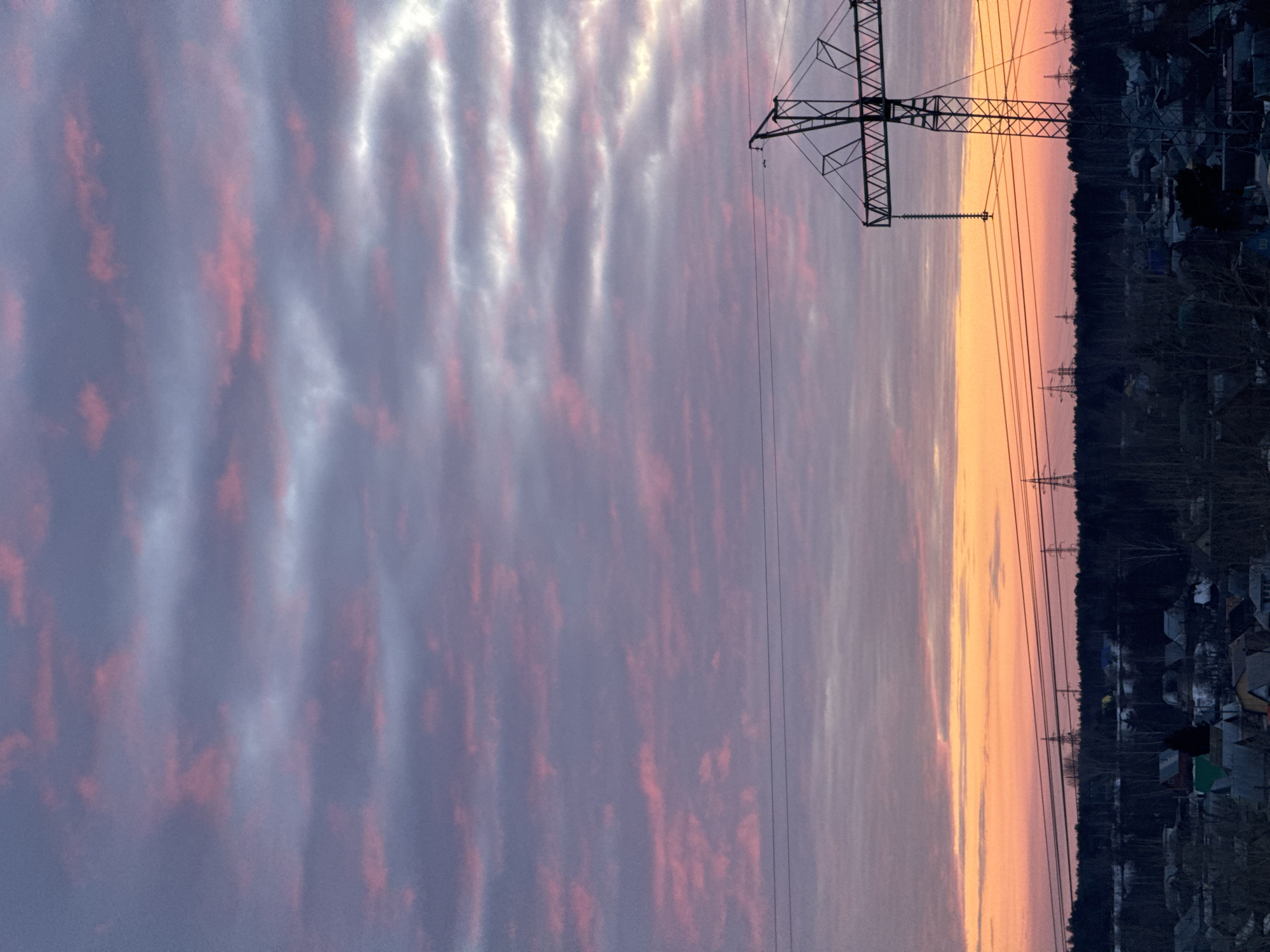 This image depicts an outdoor landscape scene during dusk or dawn in Chaikovsky, Russia, characterized by a vibrant sky and a mix of residential and natural environments.
The upper portion of the frame is dominated by a cloudy sky. Towards the zenith, the clouds are predominantly grayish-purple with scattered streaks of muted pink. As the eye moves lower towards the horizon, the sky transitions into intense orange and pink hues, indicative of the sun being below the horizon.
In the mid-ground, a dense, dark forest forms a horizontal band, silhouetted against the colorful sky. Above this treeline, multiple high-voltage power transmission lines stretch across the frame. A large, steel lattice-type power transmission tower occupies the right side of the image, with its structure extending upwards from the lower right edge and its cross-arms supporting several overhead lines that extend horizontally across the scene. Several additional, smaller power transmission towers are visible in the distance, silhouetted against the bright horizon line above the forest.
The foreground displays a residential area composed of numerous low-rise houses. These structures appear as dark silhouettes, their rooflines and architectural forms discernible. Some roofs exhibit patches of white, possibly snow. Bare deciduous trees are interspersed among the houses, suggesting a late autumn, winter, or early spring season. No individuals, active movements, or specific interactions are visible within the scene.