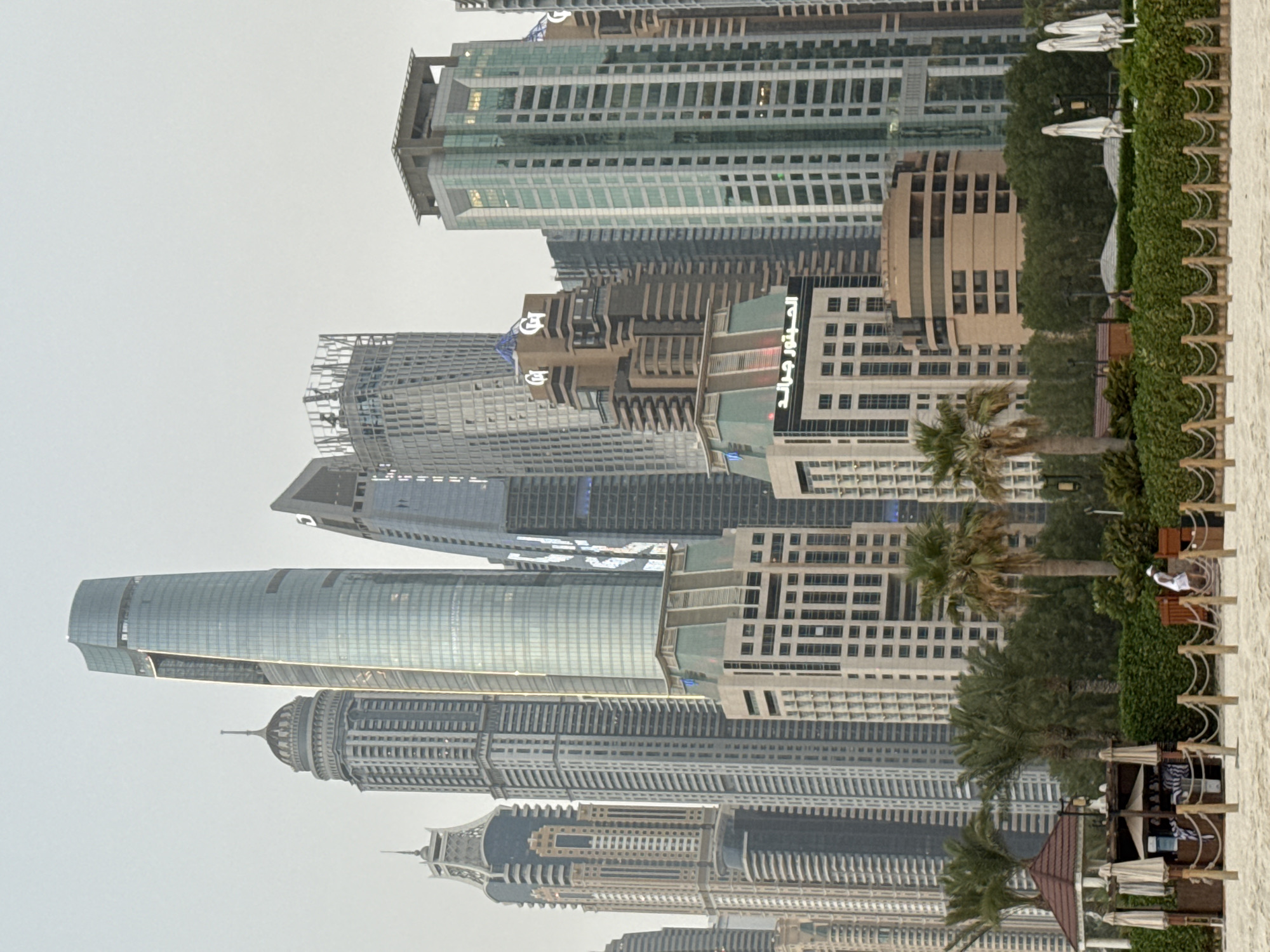 The image depicts a view of the Dubai Marina skyline in Dubai, United Arab Emirates. The foreground features a sandy beach with a low hedge and a wooden rope barrier. To the left, there's a small, open-air structure with a reddish-brown roof, housing umbrellas, chairs, and what appears to be a service counter. A person in white is visible walking on the beach towards the right. Behind the beach and vegetation, a dense cluster of modern skyscrapers dominates the background, showcasing a variety of architectural styles and heights. The sky is overcast, suggesting a hazy or diffused lighting condition.