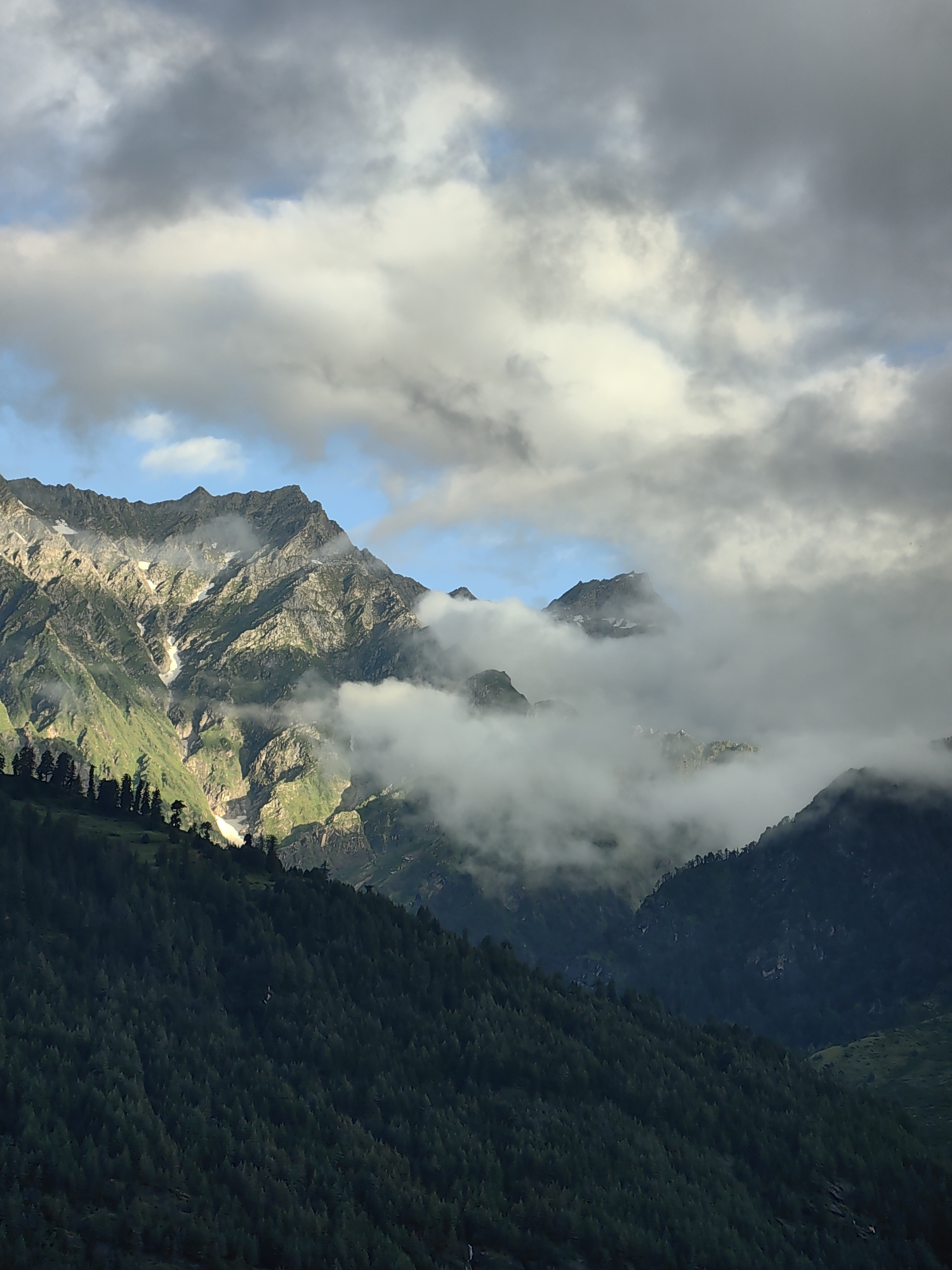 A majestic mountain landscape is depicted, featuring rugged peaks partially obscured by swirling clouds and mist. The upper portion of the image is dominated by dramatic, textured clouds in shades of grey and white, with glimpses of a blue sky peeking through. Descending from the sky, a dense layer of white mist wraps around the middle sections of the mountains, creating a sense of depth and mystery.
The mountains themselves are a mix of rocky, scree-covered slopes and areas of vibrant green vegetation, suggesting a temperate climate. Streaks of white, likely snow or ice, are visible on the higher elevations, contrasting with the darker rock and green foliage.
In the foreground, a steep, densely forested mountainside slopes downward. The trees are predominantly dark green conifers, creating a textured pattern that fills the lower part of the frame. The lighting suggests it is either early morning or late afternoon, with sunlight illuminating some of the mountain faces and creating shadows.
The scene is natural and appears to be devoid of any human presence or man-made structures, with the exception of what might be a very distant, thin line of trees along the ridgeline in the middle ground. This image captures the raw, untamed beauty of a mountainous region. Given the specific detail of Vashisht, India, this scene likely represents the breathtaking natural scenery surrounding the popular hill station, known for its hot springs and proximity to the Himalayas.