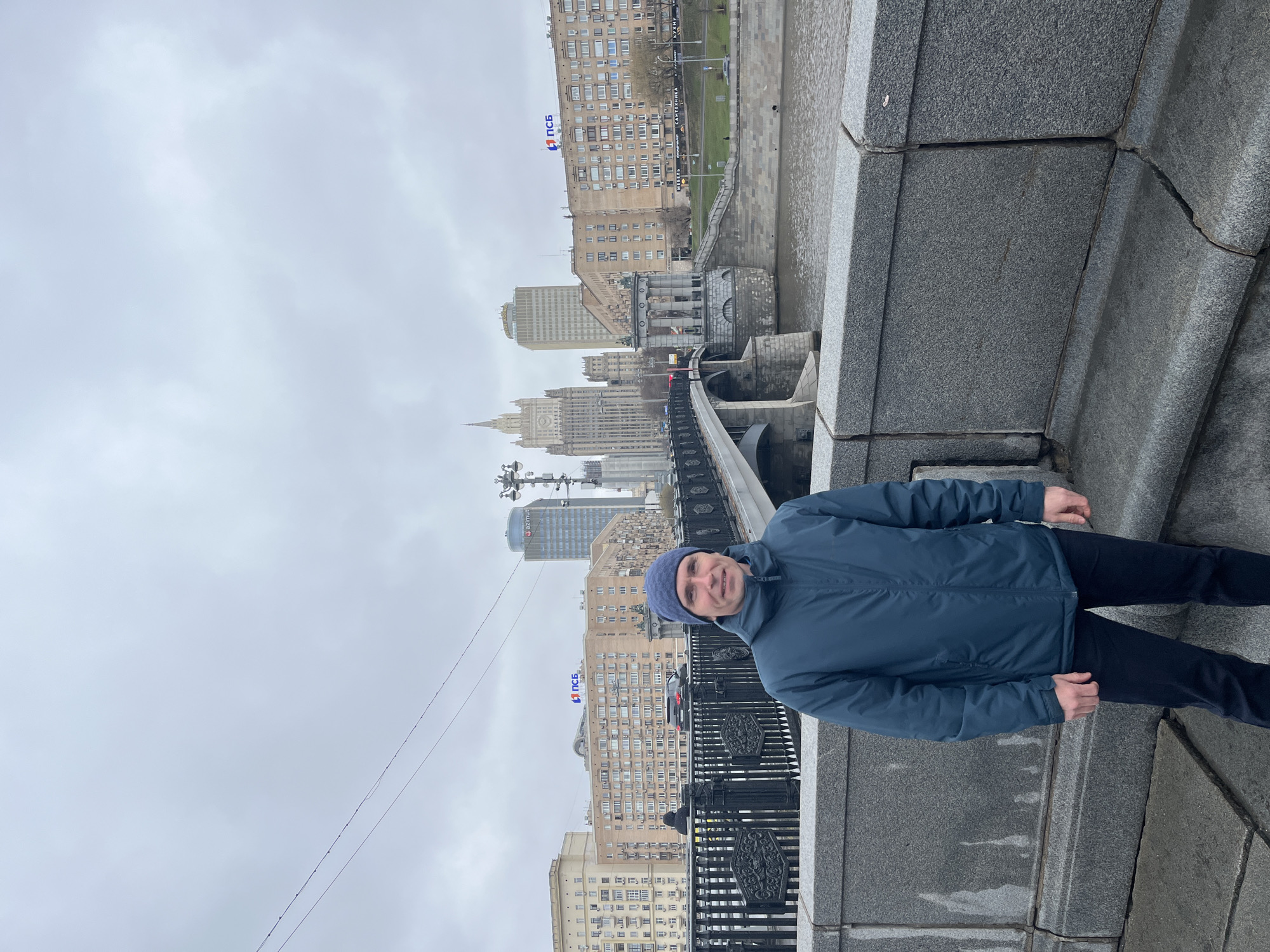 A man stands on a stone structure, likely a bridge or embankment, with a cityscape in the background. The man is wearing a blue jacket and a blue beanie. The cityscape features a mix of older and modern buildings, including the Ministry of Foreign Affairs of Russia, one of the "Seven Sisters" Stalinist skyscrapers, located in Moscow, Russia. The sky is overcast.