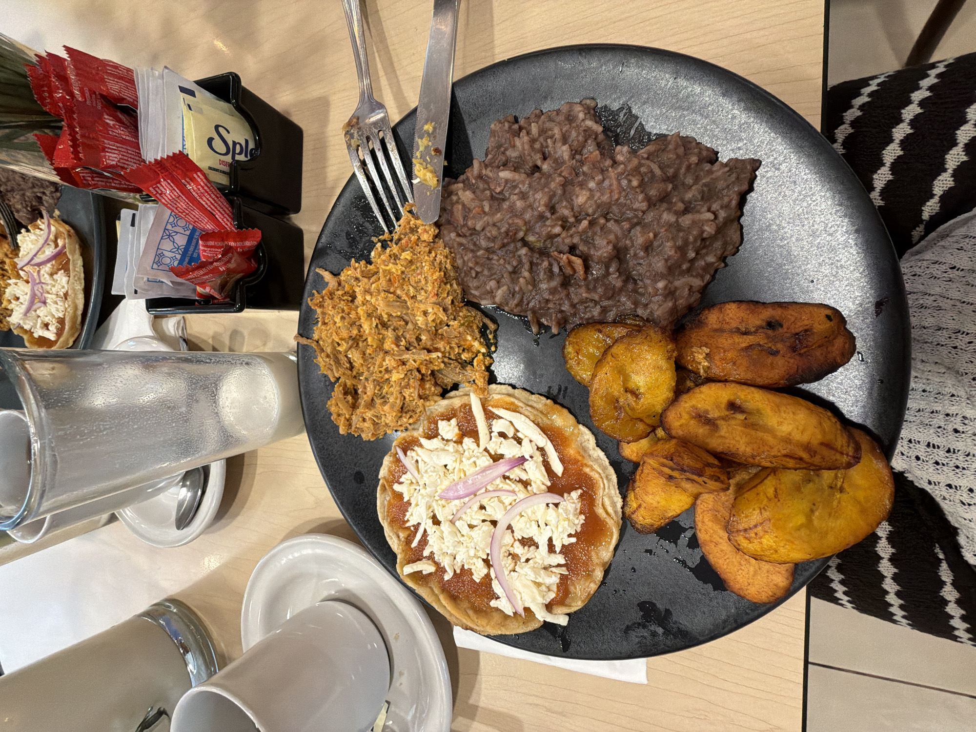 A high-angle shot presents a partially eaten meal on a black plate. Clockwise from the top, the plate contains refried beans, fried plantains (platanos), a tostada topped with what appears to be shredded cheese and red onions, and a serving of shredded meat. A fork and knife rest on the plate between the beans and the shredded meat. To the left of the plate, a small black holder contains condiment packets. Two ceramic mugs and a glass pitcher are also visible. A black and white patterned object is visible in the upper right corner, possibly a chair or cushion. The setting appears to be in a restaurant or dining area in Tuxtepec, Mexico.