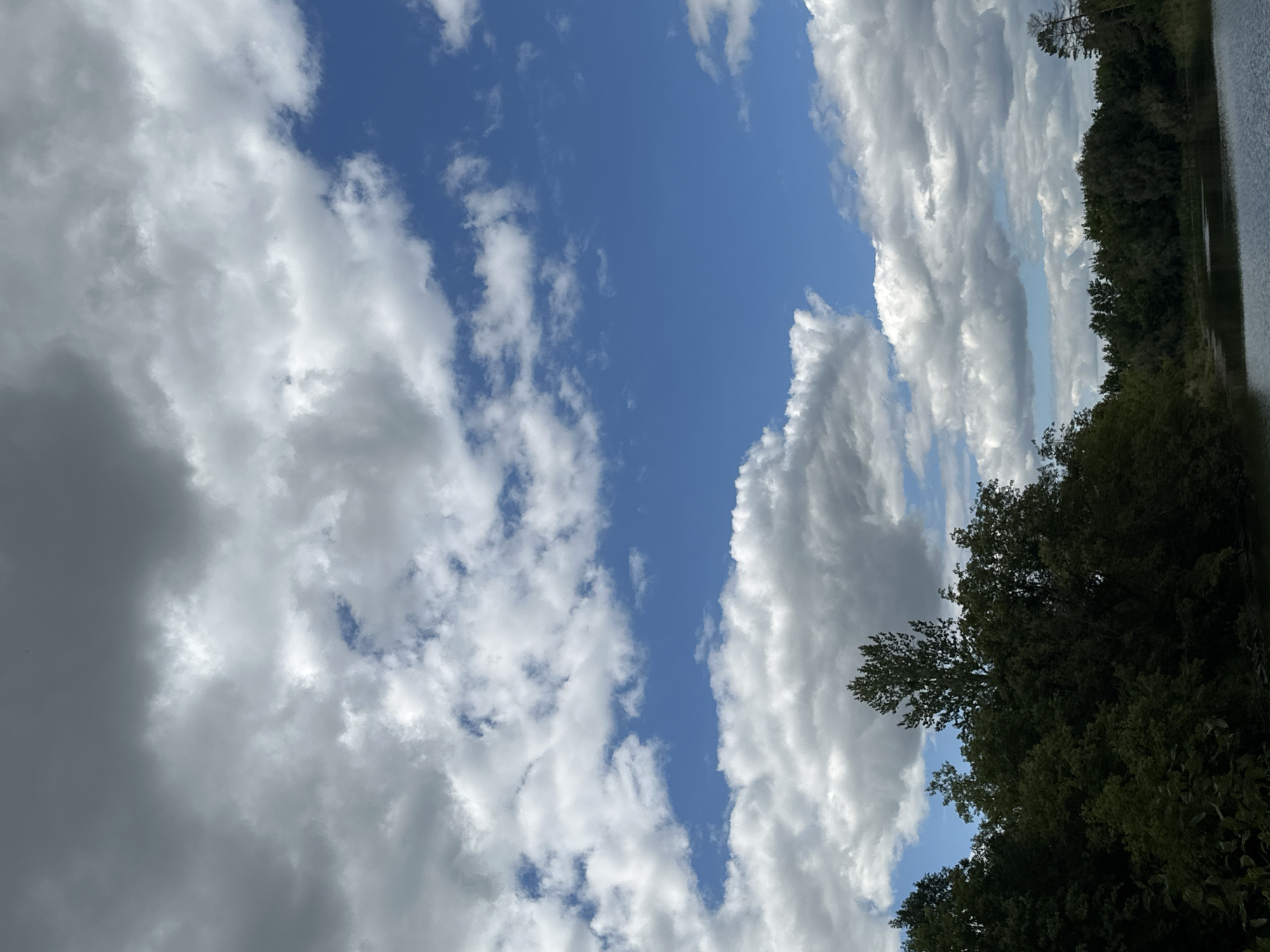 The image shows a natural landscape with a body of water in the foreground and a dense line of trees and foliage along its edge. Above the trees, a vast sky with a mix of white and gray clouds is visible against a bright blue background. The location is identified as Makiv, Ukraine. There are no people or any overt signs of human activity in the scene. The overall environment appears to be a tranquil, undeveloped area.