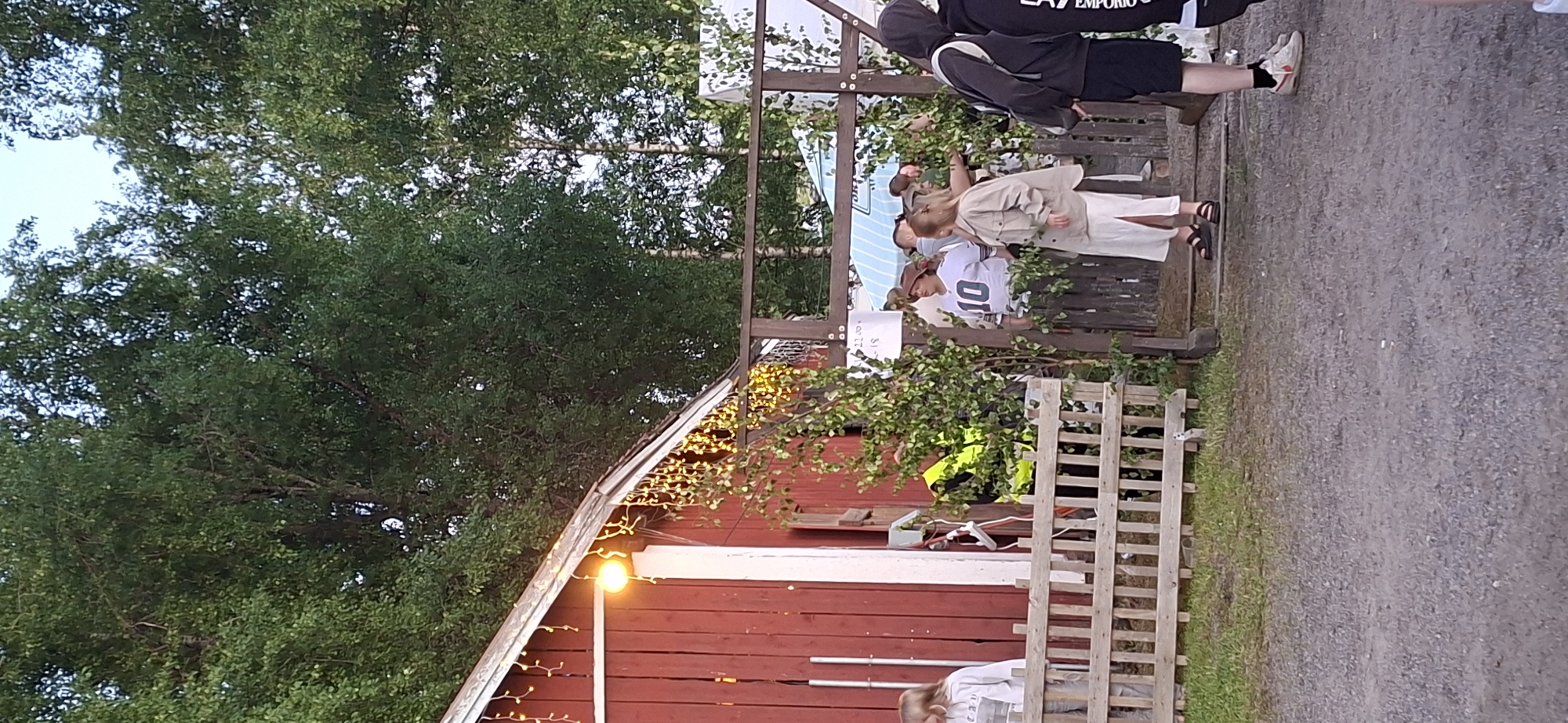 The image depicts an outdoor scene in Salpakangas, Finland, with a focus on a red wooden building adorned with decorative string lights. A group of individuals are gathered near the entrance of the building, which is accessed by wooden steps. Some people are standing, while others appear to be in conversation. The environment is characterized by a gravel pathway in the foreground and a lush green backdrop of trees. A wooden fence runs along the left side of the building. One individual is wearing a white shirt with the number 10, and another is wearing a high-visibility yellow vest. The lighting suggests it is either early morning or late evening.