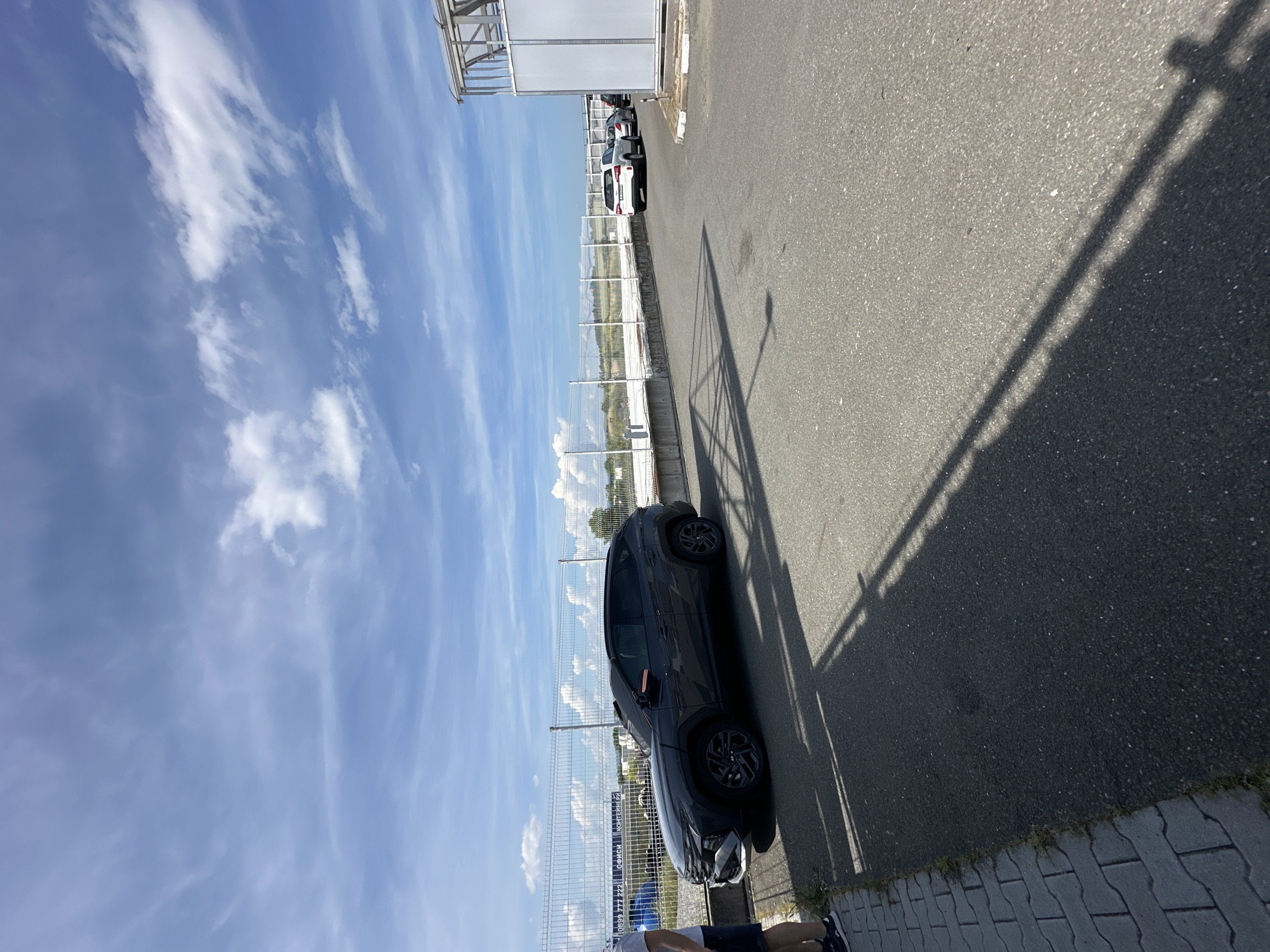 In an outdoor setting with a bright, partly cloudy blue sky, a dark gray SUV is parked on an asphalt surface. The vehicle is angled slightly towards the right. In the background, a white fence runs horizontally, with a row of other vehicles parked beyond it, including a white SUV. To the right, a modern-looking building with a white metal frame and panels is visible. A person wearing shorts and a t-shirt is partially visible on the left side of the frame, standing on a paved walkway. Shadows from the SUV and an unseen structure are cast across the asphalt. The scene appears to be an open area, possibly a parking lot or industrial space.